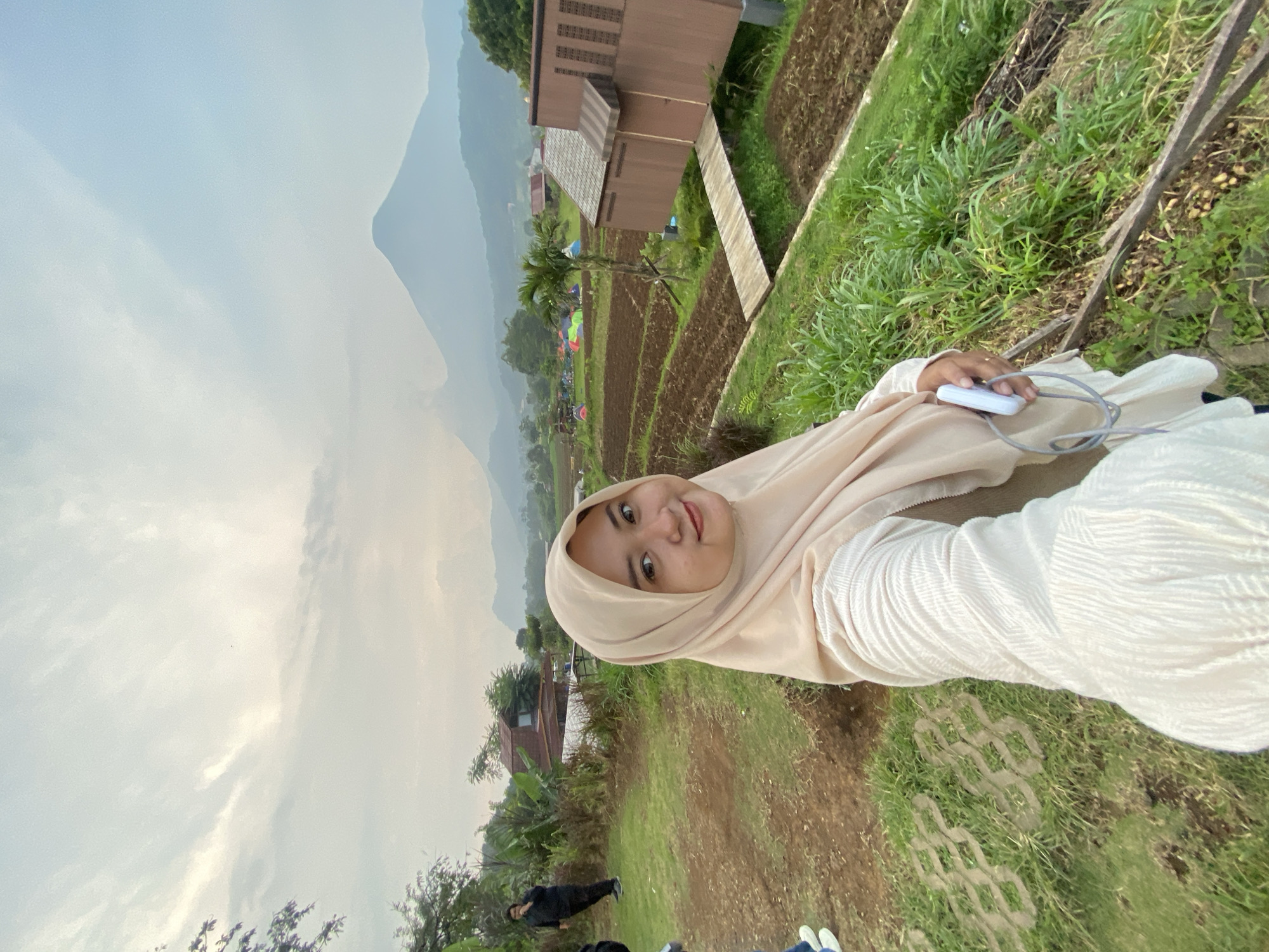A young woman in a tan hijab and white long-sleeved shirt is standing outdoors, holding a white rectangular object with a gray cord attached. She is smiling at the camera. Behind her, there are tiered rice paddies, a brown wooden building, and a mountain in the distance shrouded in mist. Tents are set up in the rice paddies. In the foreground, the ground is covered in green grass and soil. Two figures are visible in the lower part of the image. Based on visual cues, this image was most likely taken in Tamiajeng, Indonesia.