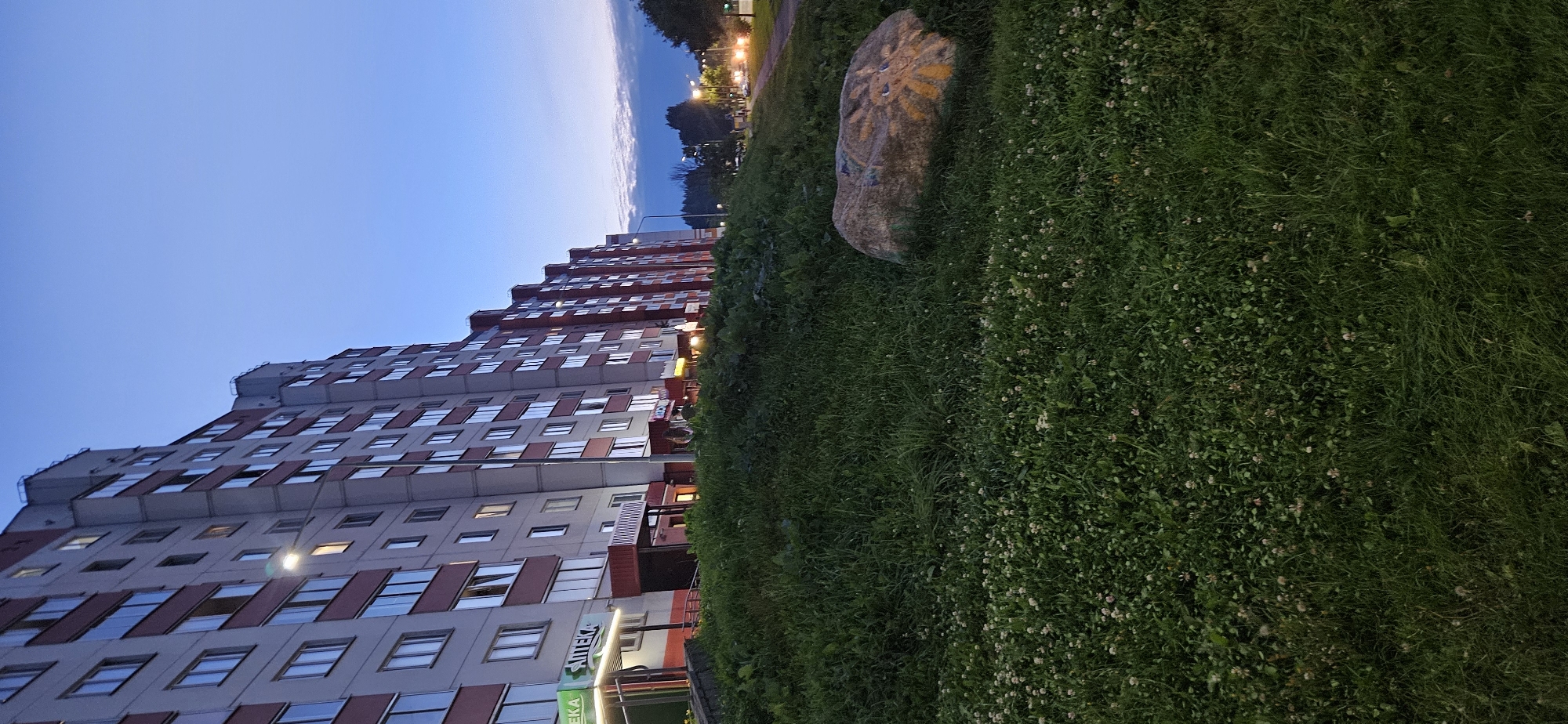 The image depicts a view of a residential building in Vitsebsk, Belarus, during dusk. The building is a multi-story structure with numerous windows, some of which are illuminated. The facade of the building is light gray with red accents. In front of the building, there is a grassy embankment with a large rock painted with a yellow sun-like symbol. A street with cars and streetlights is visible in the background. The sky is a gradient of blue and purple, indicating the transition from day to night. There are no people visible in the image. The scene appears to be a typical urban residential area.