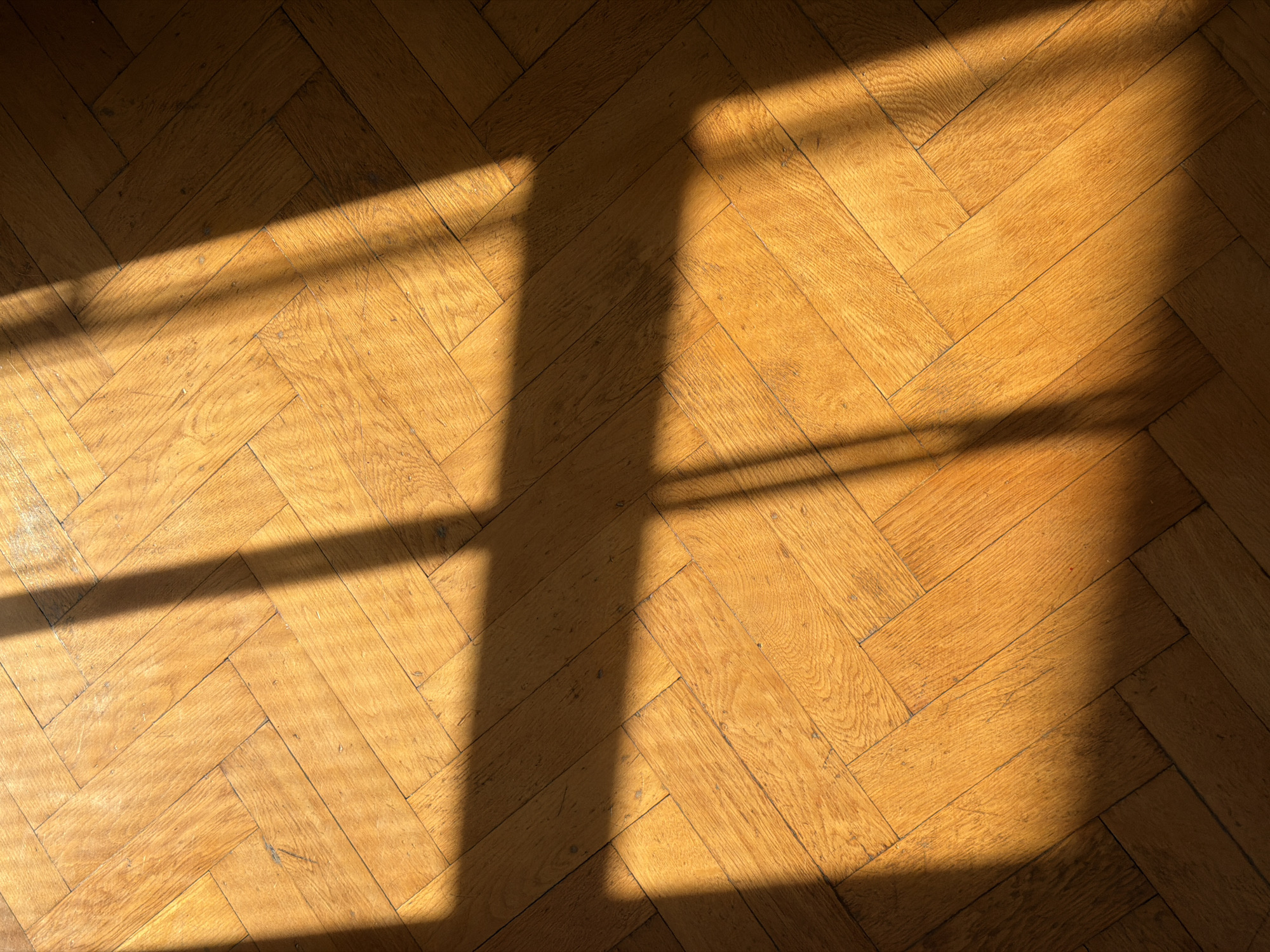 The image shows a close-up, high-angle view of a polished wooden parquet floor with a herringbone pattern. The floor is illuminated by a diagonal shaft of sunlight casting distinct shadows across its surface. The wood appears to be a warm, golden-brown color and shows signs of wear, with some minor scratches and imperfections visible. The shadows are sharp and well-defined, suggesting a strong light source, possibly from a window or skylight. The overall impression is one of a quiet, interior space bathed in natural light.