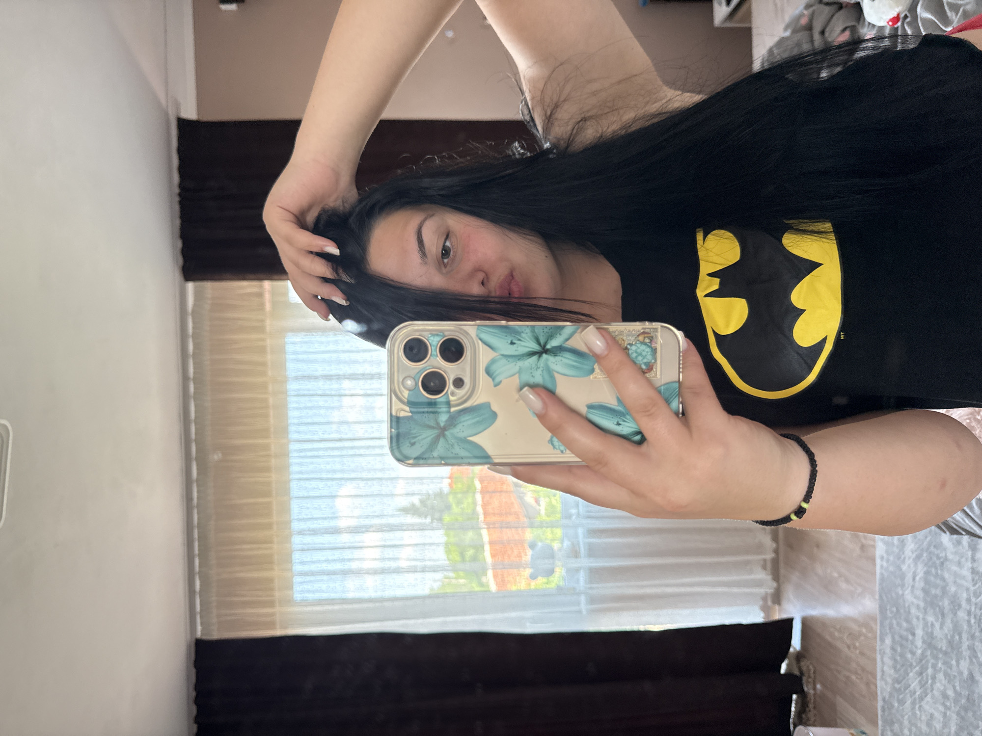 This image captures a young woman taking a mirror selfie indoors, likely in a bedroom or living room in Rogosh, Bulgaria. The image is rotated 90 degrees clockwise.
The subject is a young woman with long, dark, straight hair, dark eyes, and a fair complexion. She exhibits a neutral expression with slightly pursed lips. She is dressed in a black T-shirt adorned with a prominent yellow Batman logo, which appears reversed due to the mirror reflection. Her left hand is raised, fingers lightly touching her hair, while her right hand holds a smartphone. Her fingernails have light-colored tips, resembling a French manicure. A black beaded bracelet with a single bright green bead is worn on her right wrist.
The smartphone she holds is an iPhone, identifiable by its three-lens camera array, housed in a clear case decorated with a distinctive blue floral pattern, resembling aqua lilies.
The background features a light-colored wall. Behind the woman, a large window is visible, framed by dark brown curtains. White sheer curtains or blinds cover the window, indicating natural daylight during the day. Through these translucent curtains, glimpses of green foliage and a red-tiled roof are discernible, consistent with residential buildings in the specified location of Rogosh, Bulgaria. The overall scene conveys a casual, domestic atmosphere. No specific text is visible apart from the reversed Batman logo.