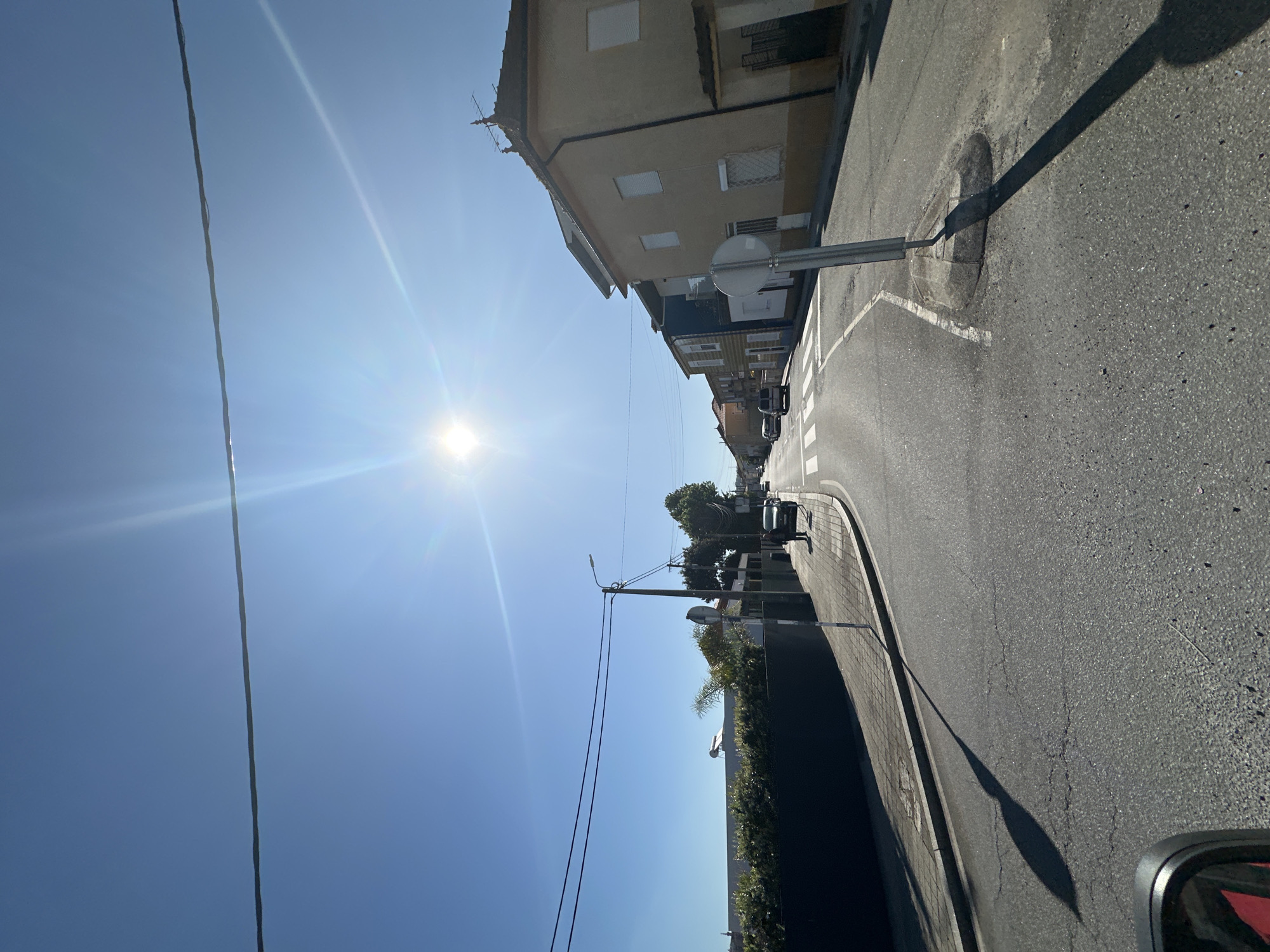 The image depicts a street scene in Gondomar, Portugal, during a bright, sunny day. The street is asphalt, with a concrete curb and sidewalk on the right. Buildings line the left side of the street, varying in height and color, but predominantly beige and tan. A traffic sign is visible on the right side of the road. Power lines run horizontally across the upper portion of the frame, intersecting with the sun's glare. A few parked cars can be seen further down the street, and some sparse vegetation is present on the side of the road. The camera seems to be positioned inside a vehicle, as indicated by a partial view of a side mirror in the lower right corner. The shadow of a light pole is visible stretching across the road.
