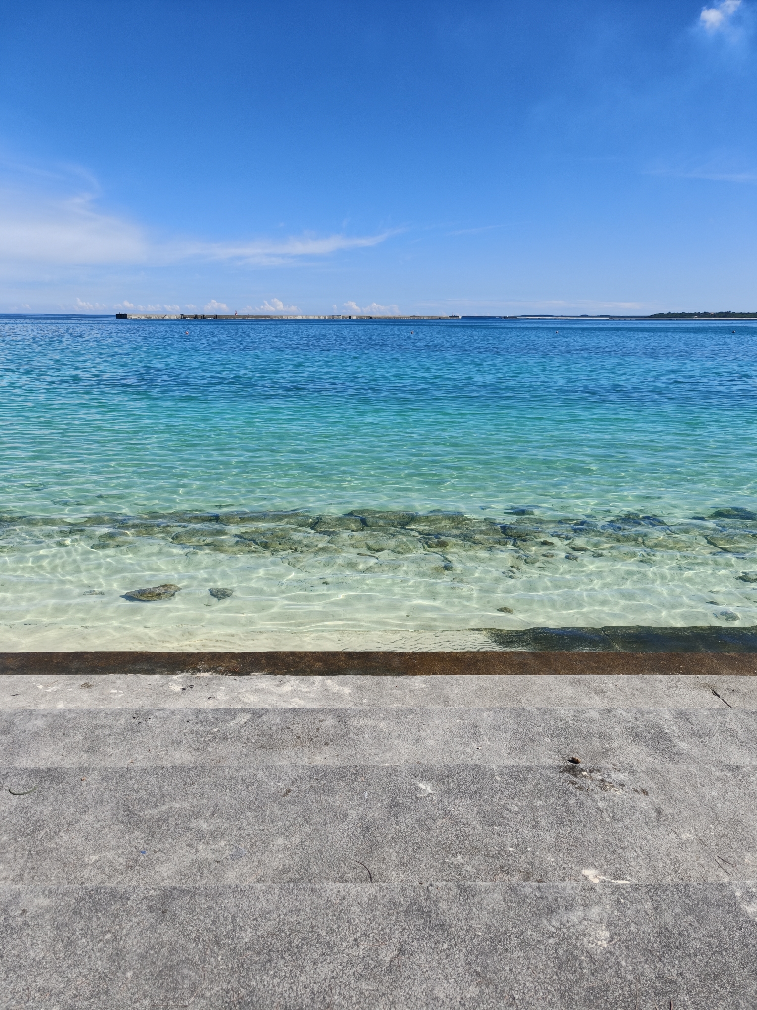This is a vertical, eye-level, medium shot of a calm, shallow, turquoise ocean in front of a concrete step structure. The water is exceptionally clear, allowing visibility of the sandy seabed and rocks. The horizon line is visible, with a breakwater or pier stretching across the middle ground, dotted with various structures, including what appears to be a lighthouse. Further in the distance, landmasses are visible. The sky is a vivid, cloudless blue. The concrete steps in the foreground are weathered and appear to be part of a seawall or access point. The scene is devoid of any people or animals. The location is likely a coastal area in Miyakojima, Japan, given the clarity of the water and the presence of such structures.