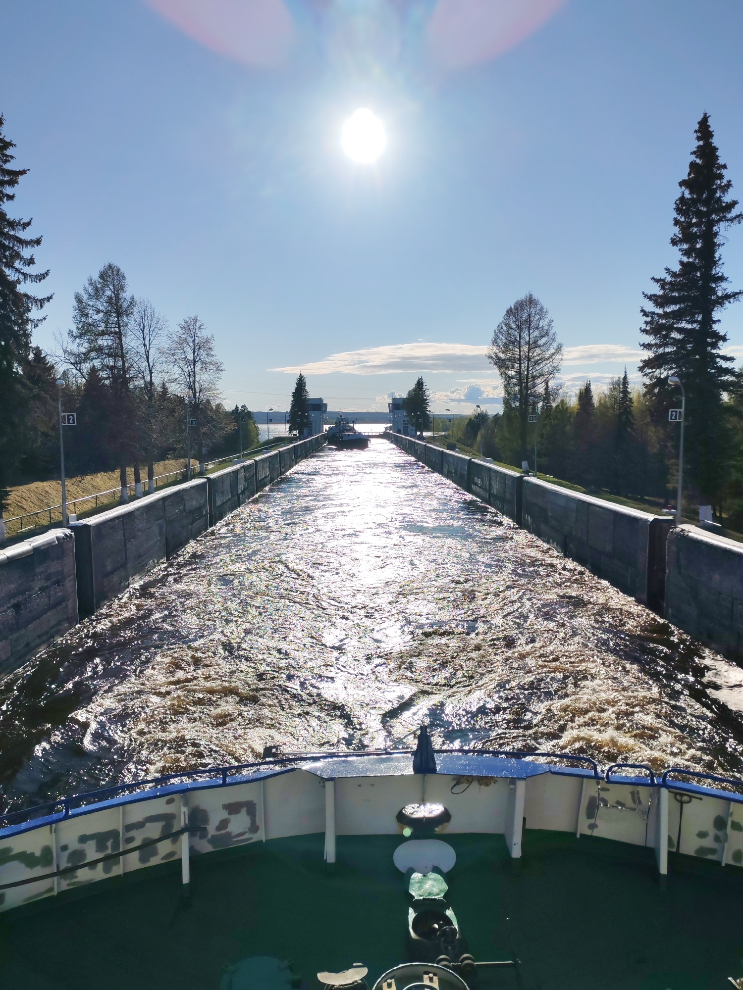 The image shows a view from the deck of a vessel traveling through a canal lock in Belousovo, Russia. The lock walls are made of concrete and appear to be slightly weathered. The water in the canal is turbulent and brown, reflecting sunlight. On either side of the lock, there are trees and vegetation. Marker posts are visible along the sides of the canal. At the end of the canal, a building and another vessel are visible. The sky is blue with the sun high in the sky.