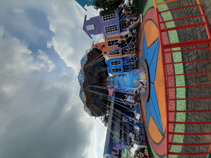 A carousel ride in a park. The carousel has a blue, orange, and green painted floor, a star-shaped design, and a metal fence around the edge. There are multiple swings on the carousel. One swing is occupied by a person in a blue shirt and jeans. The sky is partly cloudy. The carousel is located at the coordinates (-7.854, 112.485) which are in the Kecamatan Pujon area of Malang, Indonesia. The scene is likely in an amusement park or entertainment area. The image is taken from a low angle, with the bottom of the frame being almost entirely obscured by the fence. There are buildings in the background, which are all multi-colored and appear to be part of the same complex. The buildings have visible windows and doors and are in good condition. The overall scene appears to be one of fun and entertainment.