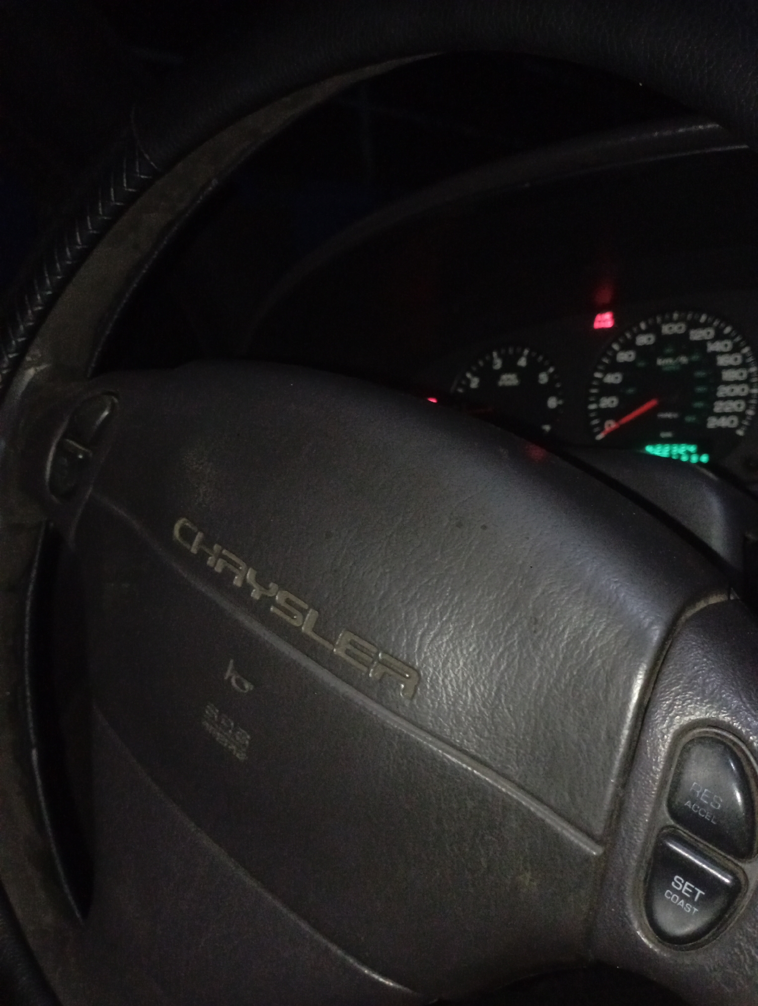 An interior shot of a vehicle, taken from the driver's perspective in Kurganinsk, Russia, depicts a dark environment with the instrument cluster illuminated. The central subject is a black Chrysler steering wheel, featuring the "CHRYSLER" emblem embossed in the center. Below the emblem, "SRS AIRBAG" is visible, along with a horn symbol. The right spoke of the steering wheel contains two control buttons: "RES ACCEL" and "SET COAST." The steering wheel rim, partially visible on the left, exhibits a stitched texture. In the background, the vehicle's illuminated dashboard instrument cluster is visible, displaying a speedometer marked up to 240 km/h with its needle at zero, and a tachometer marked up to 8 (x1000 RPM) also at zero. A red warning light is illuminated above the speedometer, and another red warning light is illuminated to the left of the tachometer. A green digital display below the speedometer shows "48754" and additional obscured green digits. No individuals are visible in the frame.