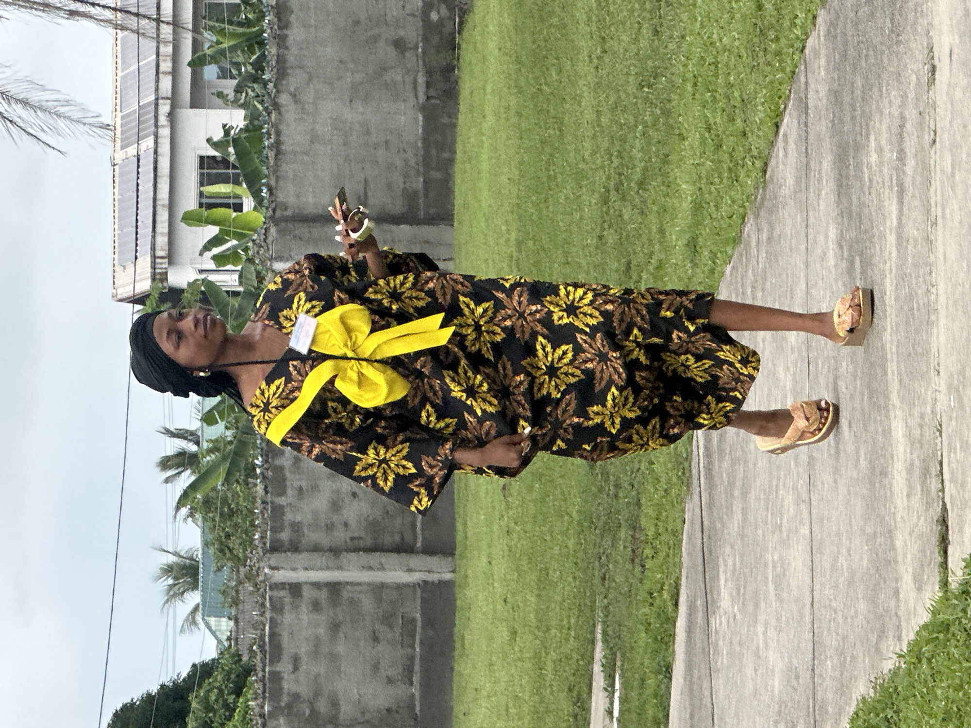 A Black woman wearing a black headscarf, a black and yellow floral-print dress, and brown platform sandals walks along a paved walkway in Itoga, Nigeria. She is looking to her left. The woman holds a smartphone in her right hand and a white bracelet is on her left wrist. Behind her is a concrete wall, a well-maintained grassy area, and a white building with solar panels on its roof. The sky is overcast.