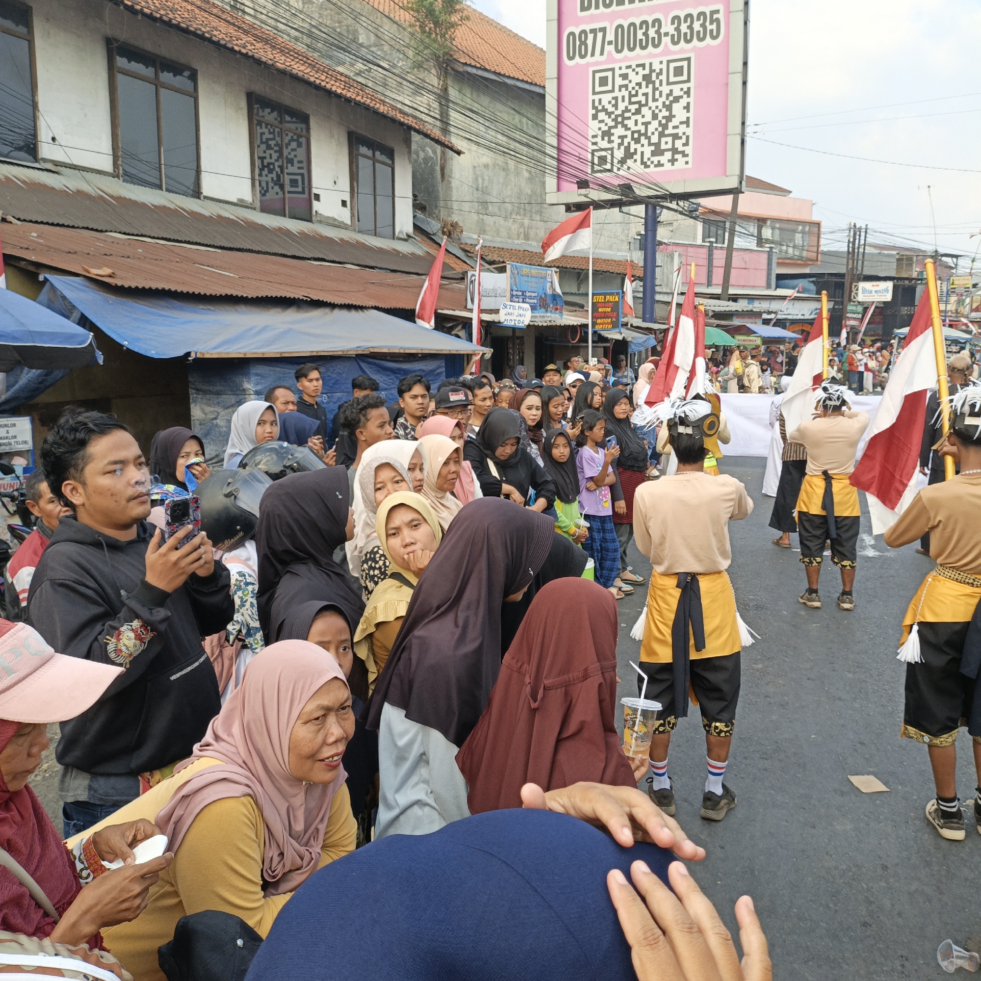 In Randudongkal, Indonesia, a lively street scene unfolds with a crowd gathered to witness a procession or performance. The atmosphere is festive, with numerous Indonesian flags, red and white, prominently displayed on poles. In the foreground, several individuals are captured in candid moments, some smoking and holding cell phones, others engaged in conversation, and children observing the proceedings.
The architecture visible includes buildings with tin roofs and older facades, suggesting a traditional urban environment. A prominent billboard with a QR code and phone number is visible in the background, indicating commercial activity.
The central focus of the scene includes performers in traditional attire, wearing distinctive headwear adorned with white feathers and elaborate waist sashes. They are holding flags and appear to be part of a cultural display or parade. The ground is paved with asphalt, and the sky is overcast. The presence of tents and makeshift stalls hints at a market or event taking place. The overall impression is one of community gathering and cultural celebration.