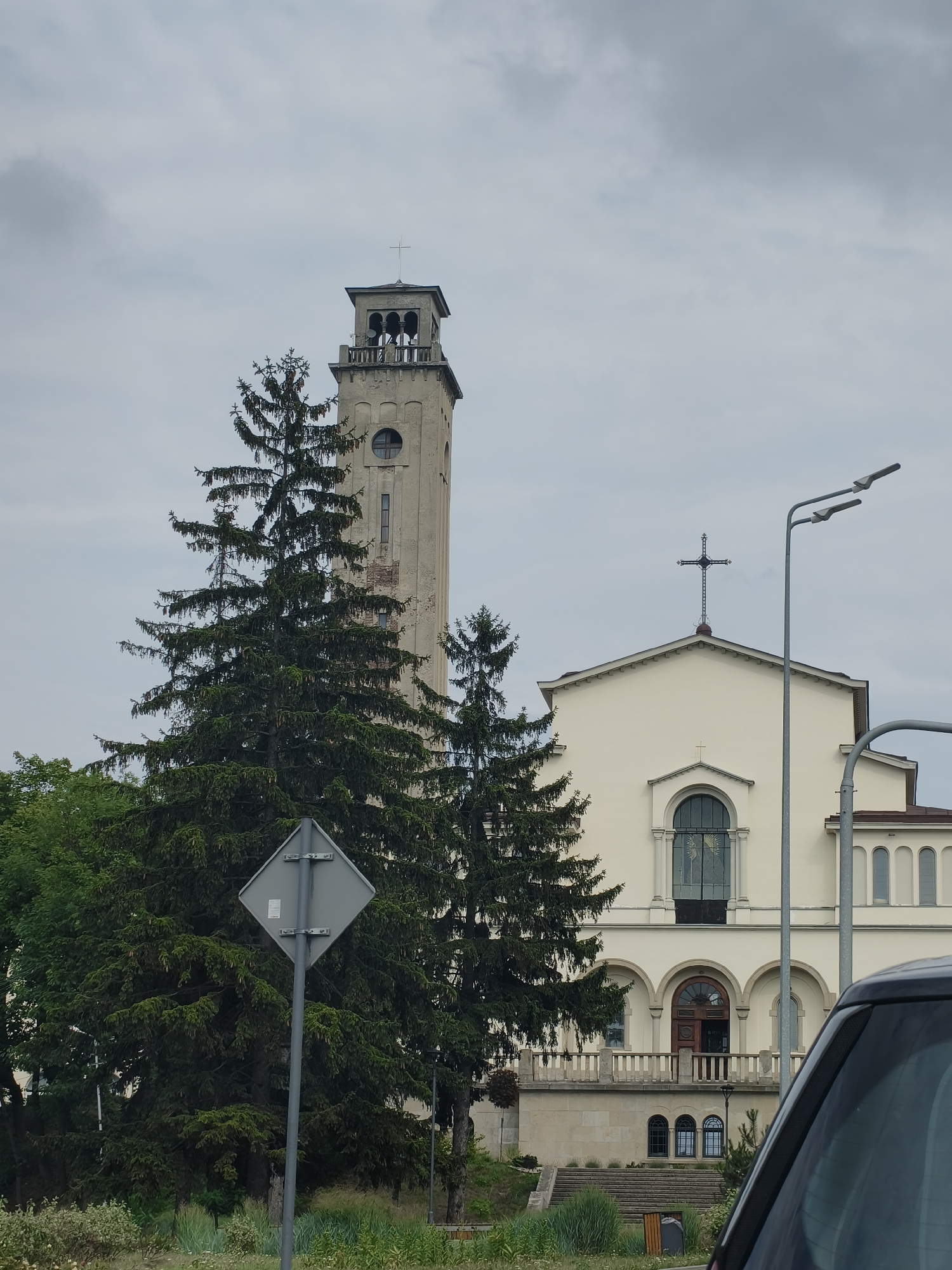 The image captures a church in Lviv, Ukraine. The church features a tall, slender bell tower with arched openings near the top, a circular window and a rectangular window on its facade. The main building of the church is cream-colored with arched windows and a balcony with a railing. A prominent cross is visible on the roof of the church. Large pine trees are situated in front of the church, obscuring parts of its structure. A street sign is positioned in the foreground to the left, and the corner of a dark-colored car is visible on the right side of the frame. The sky is overcast.