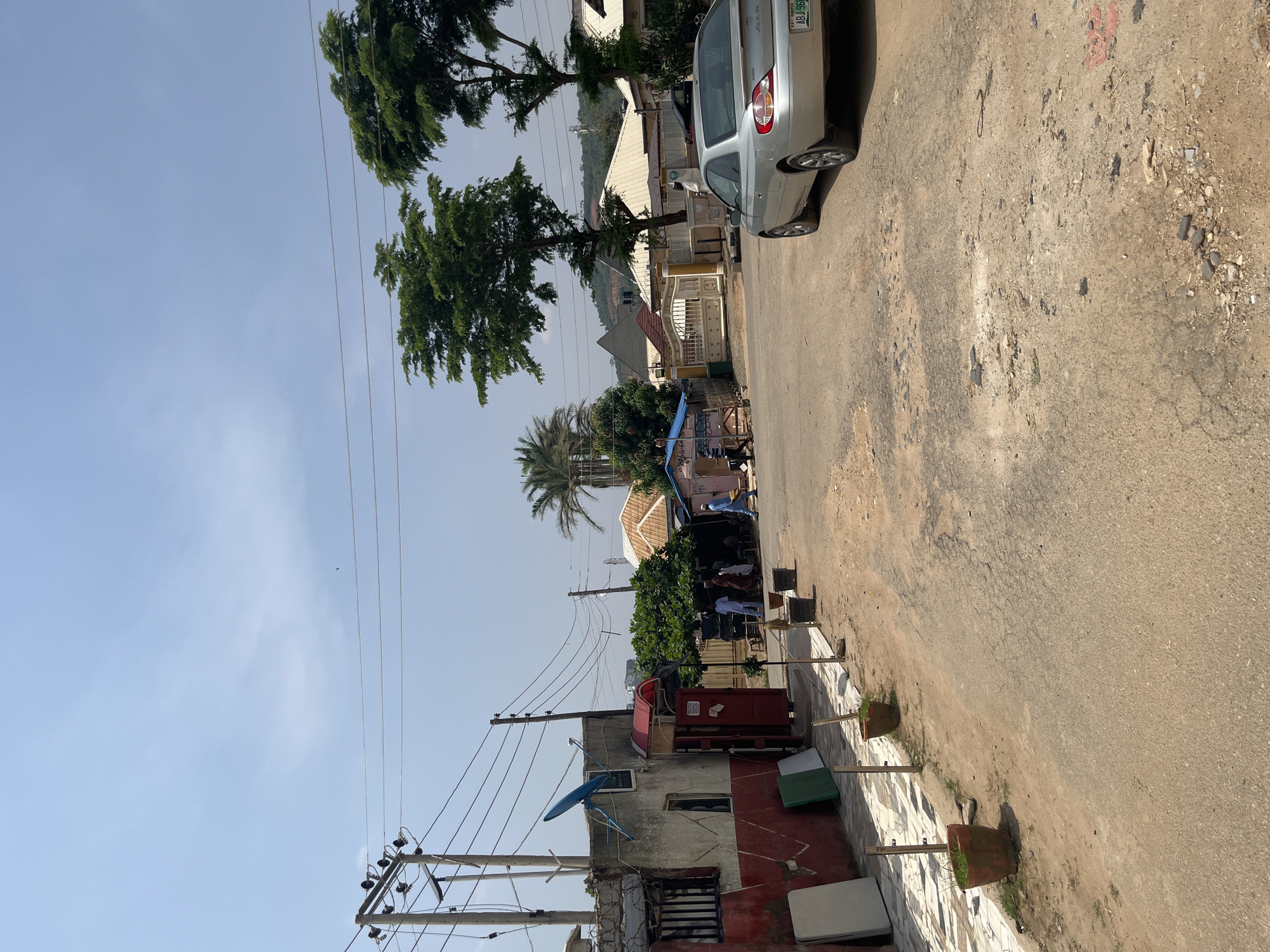 This is a sideways photo taken outdoors on a sunny day in Abuja, Nigeria. The scene depicts a street with buildings, trees, power lines, and a silver car parked on the right side of the road.
In the foreground, a dirt and asphalt road is visible, with some debris scattered on it. Several potted plants are lined up along the edge of the road. Behind the plants, there are buildings with red and white painted walls. Power lines and poles are prominent throughout the image, with a satellite dish attached to one of the buildings.
Further back, more buildings with various roof styles are visible, interspersed with lush green trees. A silver car is parked on the right side of the road, facing away from the viewer. The license plate reads "ABJ 966 J." Some people appear to be gathered in front of one of the buildings, under a canopy. A small bird is visible in the sky.
The overall impression is of a typical street scene in a residential or commercial area in Nigeria, on a clear and bright day. The sunlight casts shadows, suggesting it is likely midday or early afternoon.