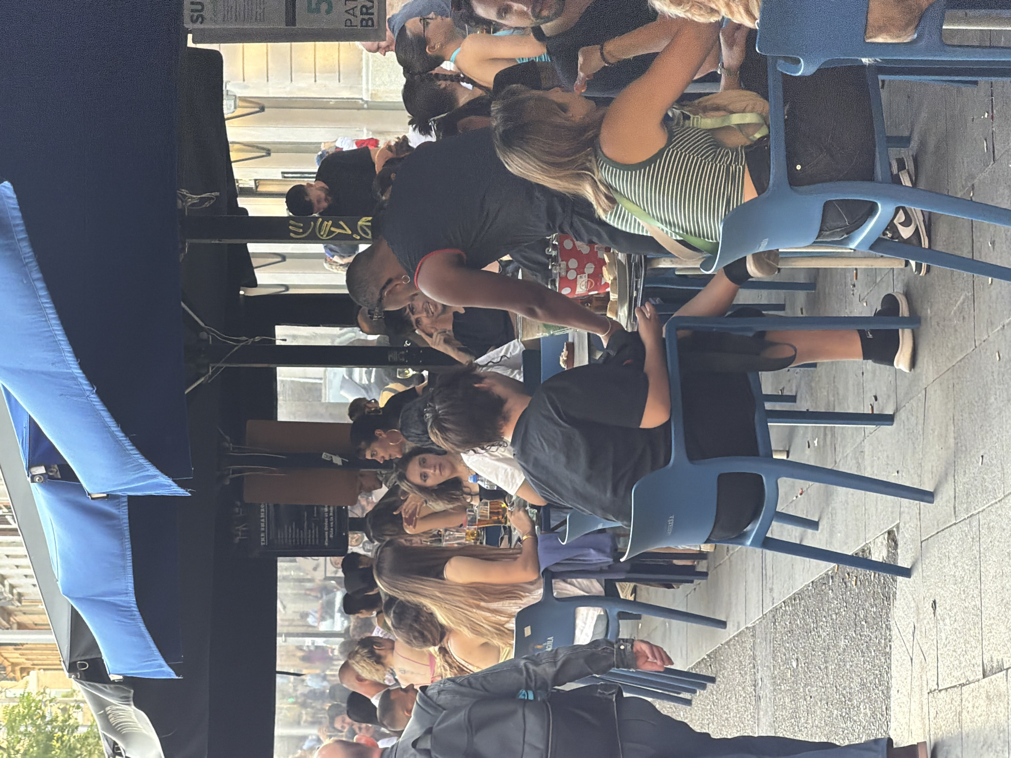 The image depicts an outdoor seating area of a restaurant or bar, likely located on a pedestrian street in Barcelona, Spain. The scene is bustling with people seated at tables under a large blue awning. In the foreground, a person in a black t-shirt and shorts walks away from the camera, wearing a backpack. Several other individuals are seated at tables, some are eating or drinking, and others are engaged in conversation. The tables are set with plates, glasses, and bottles of drinks. The environment appears to be a warm, sunny day, with buildings visible in the background. The overall atmosphere is casual and social, with patrons enjoying the outdoor ambiance.