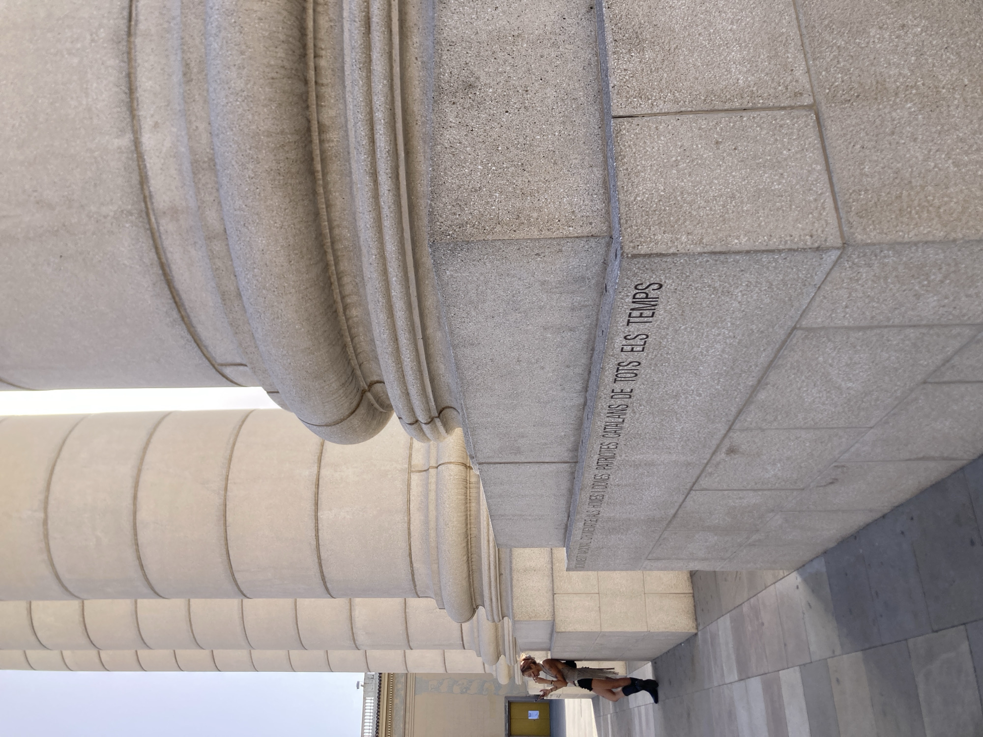The image captures a close-up view of monumental stone columns, characteristic of classical architecture, likely in an outdoor urban setting. A young woman, dressed in dark shorts and boots with a light-colored top and a headband, is leaning against the leftmost column and looking at her phone. She has a backpack slung over her shoulder. The columns are massive, constructed from light-colored stone, with distinct horizontal banding. An inscription is visible on the stone base of one of the columns. The ground in front of the columns is paved with large rectangular tiles, laid in a grid pattern. In the background, a building with a yellow facade and a yellow door can be seen. The lighting suggests a bright, sunny day. The inscription on the column base reads in Catalan: "AQUEST MONUMENT DEDICAT ALS HOMES I DONES PATRIOTES CATALANS DE TOTS ELS TEMPS". This monument is located at the Palau Nacional in Barcelona, Spain, a landmark building that houses the Museu Nacional d'Art de Catalunya (MNAC).