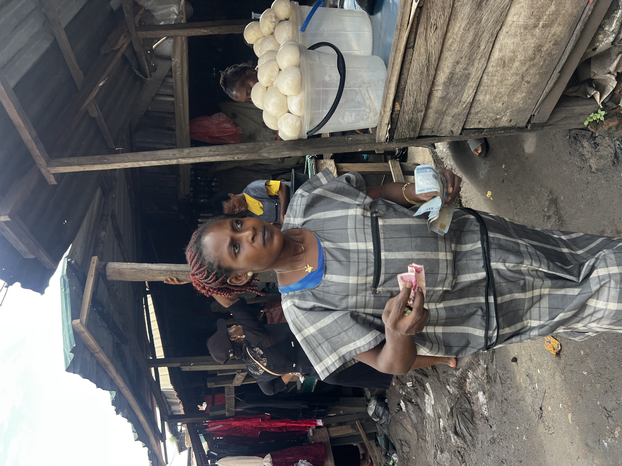 The image depicts a street market scene in Nkpor, Nigeria. In the foreground, a woman with braided red hair, wearing a grey and white checkered dress and a gold necklace, holds several Nigerian naira banknotes in her hands. She appears to be engaged in a transaction. Behind her, a woman in a black top and a baseball cap is visible, facing away from the camera. In the background, two other women are present, one in a yellow and grey top and the other partially obscured.
The environment is a bustling marketplace with wooden stalls and a corrugated iron roof. Various goods, including clothing and what appear to be food items in buckets, are displayed. The ground is muddy and littered, indicating typical market conditions. The overall atmosphere is one of everyday commerce in a vibrant Nigerian urban setting.