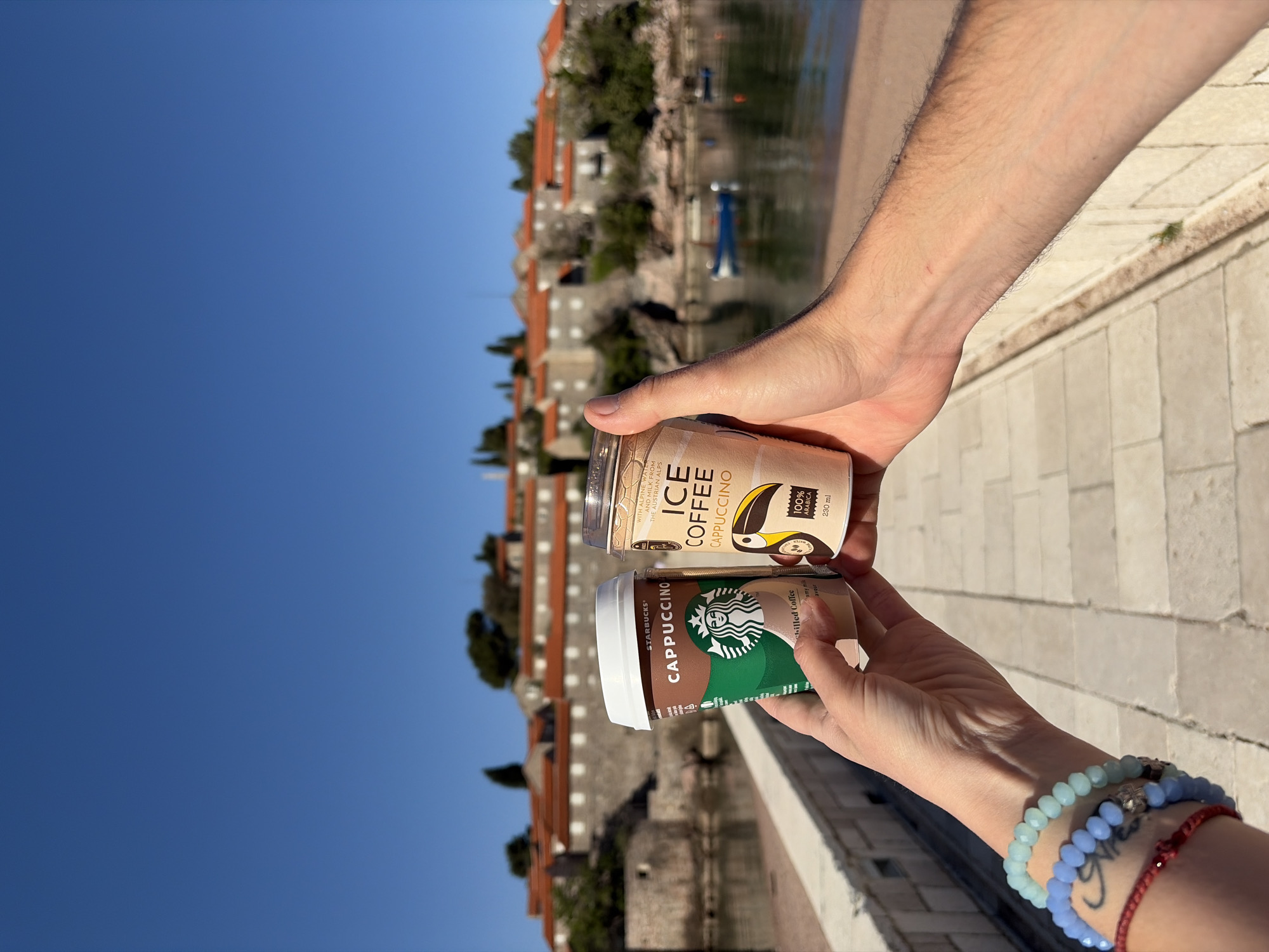 A rotated outdoor shot captures two people's hands holding ready-to-drink coffee beverages against a scenic coastal backdrop, identified as Sveti Stefan, Montenegro. The image is taken in bright daylight under a clear blue sky, suggesting sunny weather.
In the foreground, a left hand, adorned with multiple beaded bracelets in shades of light blue and darker blue, and a red string bracelet, holds a green and brown Starbucks Cappuccino can. Adjacent to it, a right hand holds a beige can of "ICE COFFEE CAPPUCCINO." The labels on the cans are clearly visible, with the Starbucks can featuring its signature logo and "CAPPUCCINO" text. The other can prominently displays "ICE COFFEE CAPPUCCINO," with additional text noting "WITH ALPINE MILK FROM THE AUSTRIAN ALPS," "100% ARABICA," and "230 ml," alongside "chilled Coffee" and "flavour."
The background reveals a picturesque coastal village with traditional stone buildings featuring distinctive orange-tiled roofs. These structures line the shore of a body of water, likely the Adriatic Sea. Small, indistinguishable boats can be seen on the water. The setting implies a leisure activity, possibly a coffee break while enjoying the historic and beautiful views of Sveti Stefan.