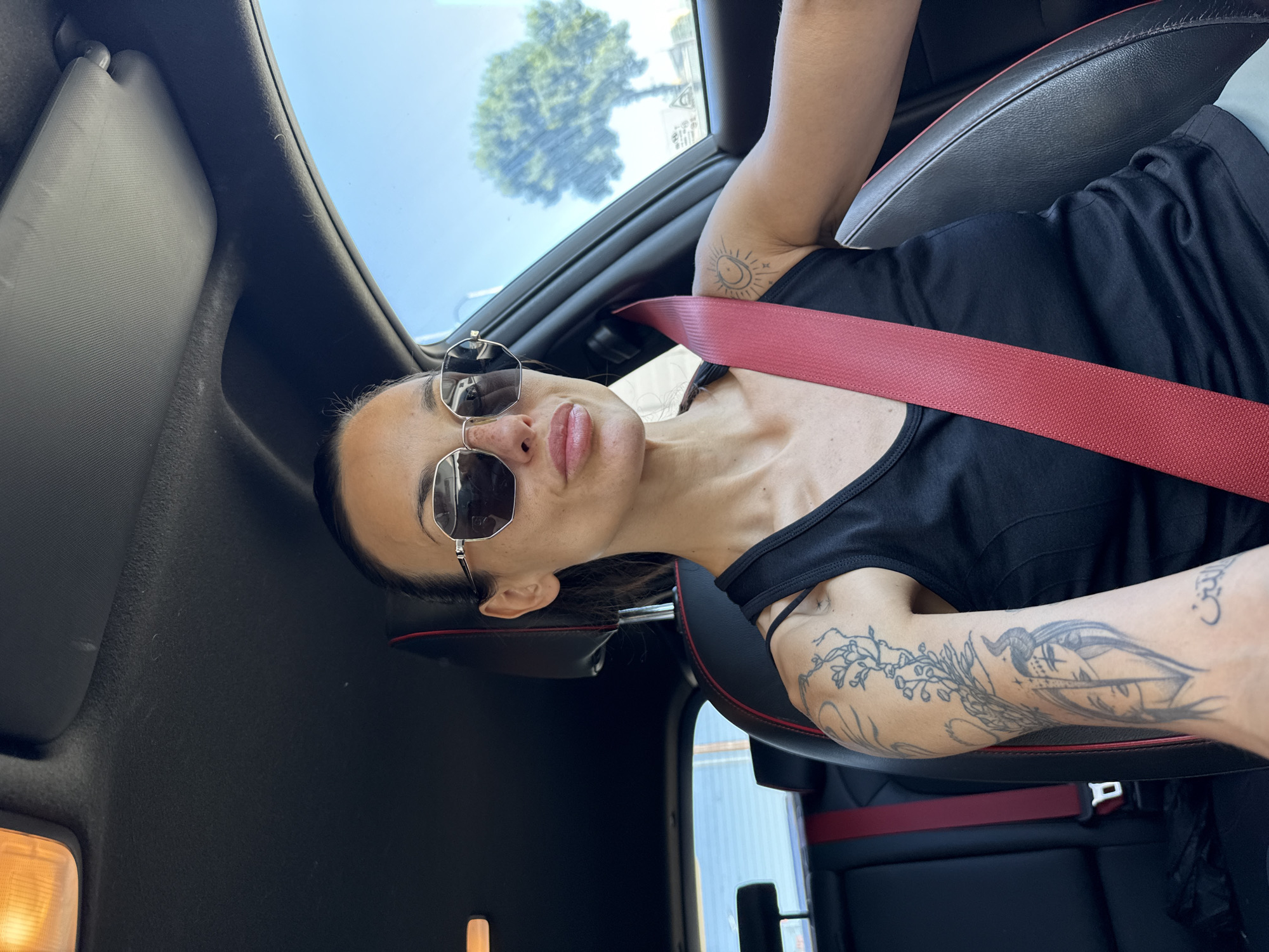 This media file shows a woman sitting in the passenger seat of a car, likely a convertible given the open sunroof. She is wearing a black tank top and sunglasses. Her left arm is tattooed with several designs, including a sun and moon, and what appears to be a face with horns. A red seatbelt is visible across her chest. The car's interior is dark, with black leather seats accented with red trim. Through the sunroof, a bright blue sky and a tree are visible, suggesting it is daytime and likely good weather. The woman's expression is neutral. There is no visible text in the image. The setting is a car, and the activity is simply sitting in the car, possibly on a drive. The overall impression is one of casual leisure.