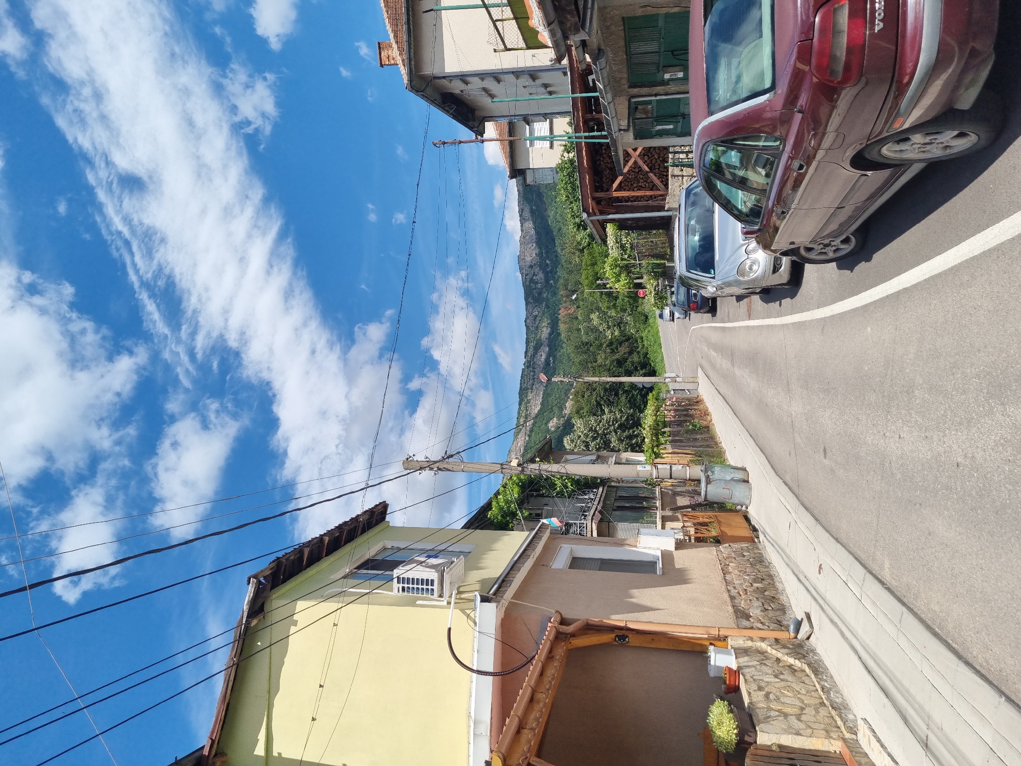 This image captures a street scene in the city of Belogradchik, Bulgaria, under a partly cloudy blue sky. A paved road with a white lane marking runs diagonally through the frame, curving to the right. On the left side of the road, a two-story building with a pale yellow facade and a brown tiled roof is visible. This building features windows, an air conditioning unit mounted near a window, and a covered porch area with wooden beams. Further down the street on the left, a Bulgarian flag is displayed on a balcony.
On the right side of the road, a series of buildings are present. The closest building has a light-colored facade, a red tiled roof with a chimney, and an attached structure with green doors. Next to this is another building with a more weathered facade and a covered area piled with firewood. Beyond that, additional houses and fences line the street.
Several cars are parked along the right side of the road. Closest to the viewer is a maroon Volvo sedan. Further down the road, a silver sedan, a black sedan, and a white car are parked in a row. In the background, the road continues, flanked by vegetation and leading towards a scenic mountain range with a rugged, rocky face and lush green slopes. Utility poles with numerous wires are prominent throughout the scene, indicating active infrastructure. The overall impression is of a residential street in a town situated in a hilly or mountainous region.