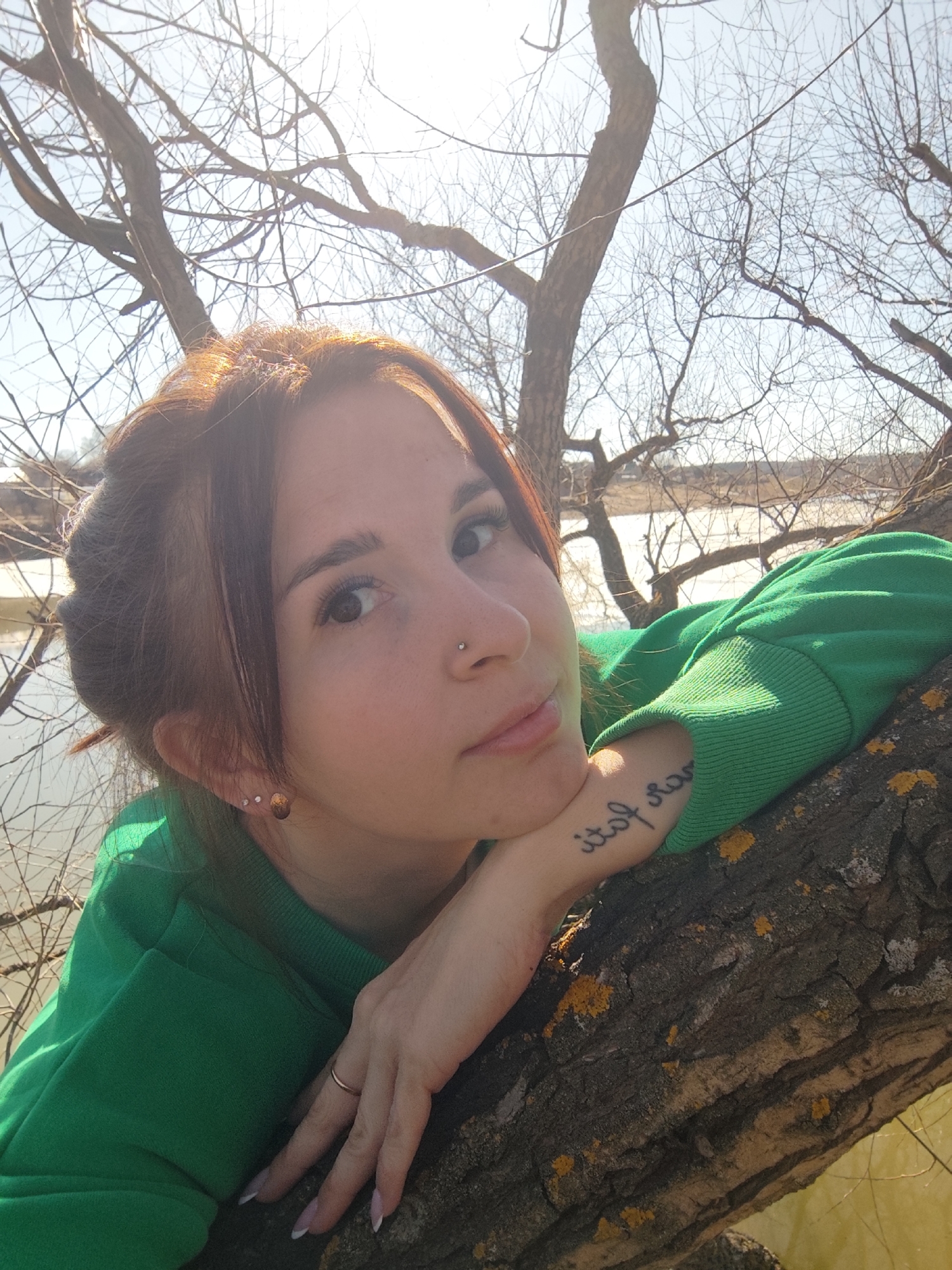 A young woman with reddish-brown hair is featured in a close-up, outdoor portrait taken in Фоки, Russia, during bright daylight. Her hair is styled with some strands framing her face, glistening in the sunlight. She has a small silver stud nose piercing on her right nostril and three small stud earrings in her left ear. She wears a vibrant green sweatshirt.
The woman is posing for the camera, resting her chin on her left hand, which is supported by her left forearm. Her arm is draped over a thick, gnarled tree branch that horizontally traverses the midground. The tree's bark is rugged and textured, adorned with patches of yellow-orange lichen. A simple gold-toned ring is visible on her left ring finger, and her fingernails display a French manicure. A cursive script tattoo is visible on her inner left forearm; it appears reversed in the image, with fragmented letters resembling "itof sa" when read directly.
She looks directly at the camera with a calm and gentle expression, possibly thoughtful or serene. In the background, numerous bare tree branches are visible, indicating a deciduous tree in late winter or early spring. Behind the trees, a body of water—likely a river or lake—reflects the bright, clear sky. The scene conveys a peaceful, natural setting bathed in strong sunlight.
