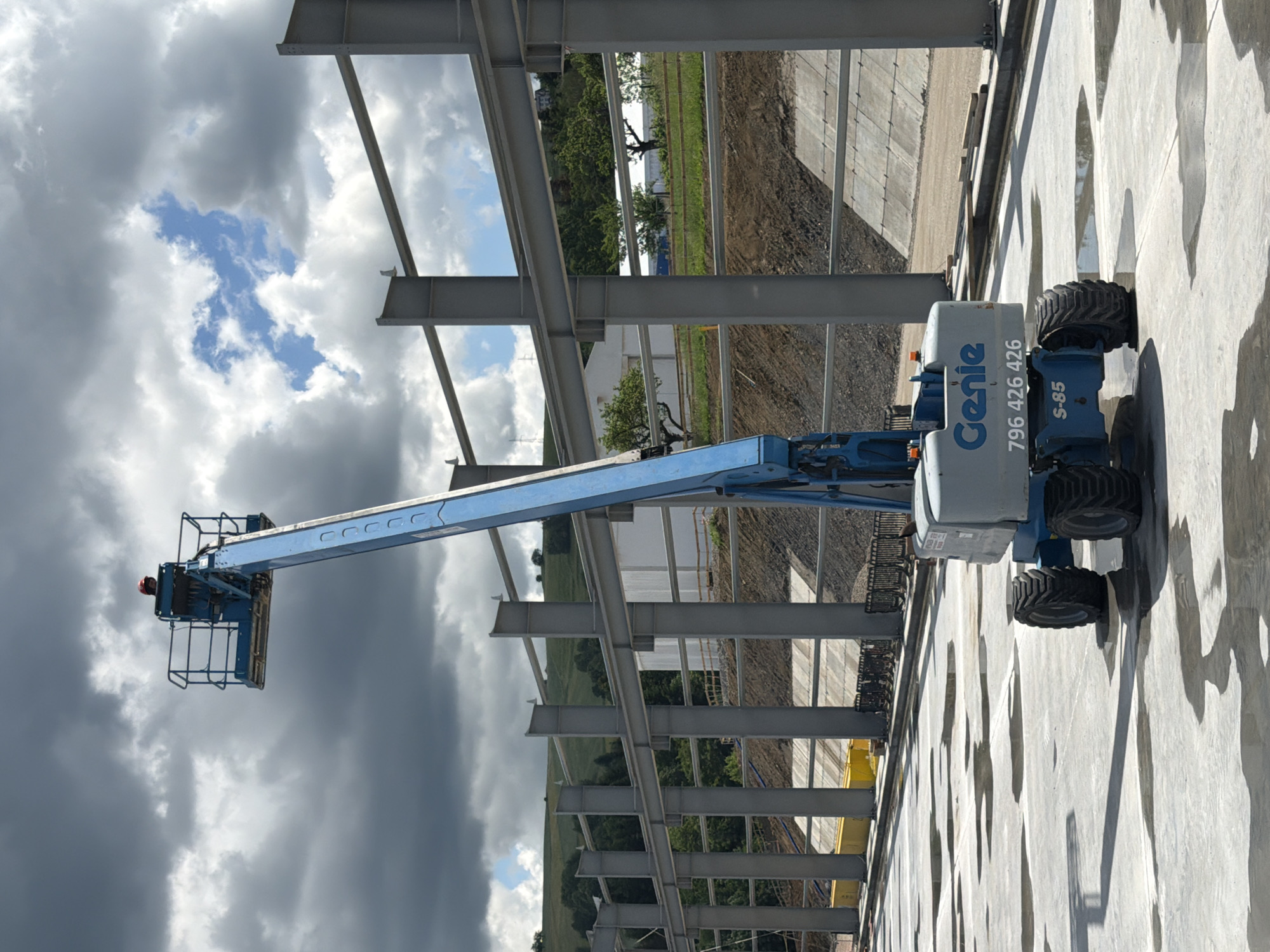 An aerial work platform, identified by the branding "Genie" and model number "S-85", is positioned on a wet, concrete surface. The platform is extended upwards and features a blue basket at the top, where a person wearing a red hard hat is visible. In the background, construction of a large steel-framed building is underway, with massive grey steel beams and columns forming the skeletal structure. The ground is a mix of excavated earth and concrete retaining walls. The environment appears to be a construction site located in Stróża, Poland, under a dramatic sky with dark, heavy clouds interspersed with patches of blue.