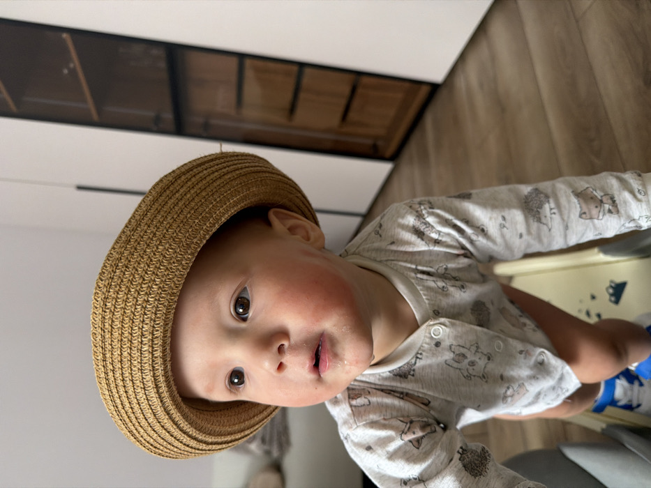 The image displays a baby or toddler in an indoor domestic environment, located in the city of Chaikovsky, Russia. The child is observed in an upright position, potentially supported by a low-lying, light-colored object in the foreground, and is looking towards the upper left. The child wears a light brown straw hat, a light grey long-sleeved onesie or top adorned with various animal patterns, and blue and white shoes. Bare legs are visible beneath the clothing. Facial features include light brown eyes, redness on both cheeks, and a small quantity of saliva on the chin and lower lip. The background consists of light-colored walls and a light brown wooden floor. To the right, a tall, dark-framed cabinet with glass doors is visible. A blurred light-colored furniture piece is partially visible in the left background.