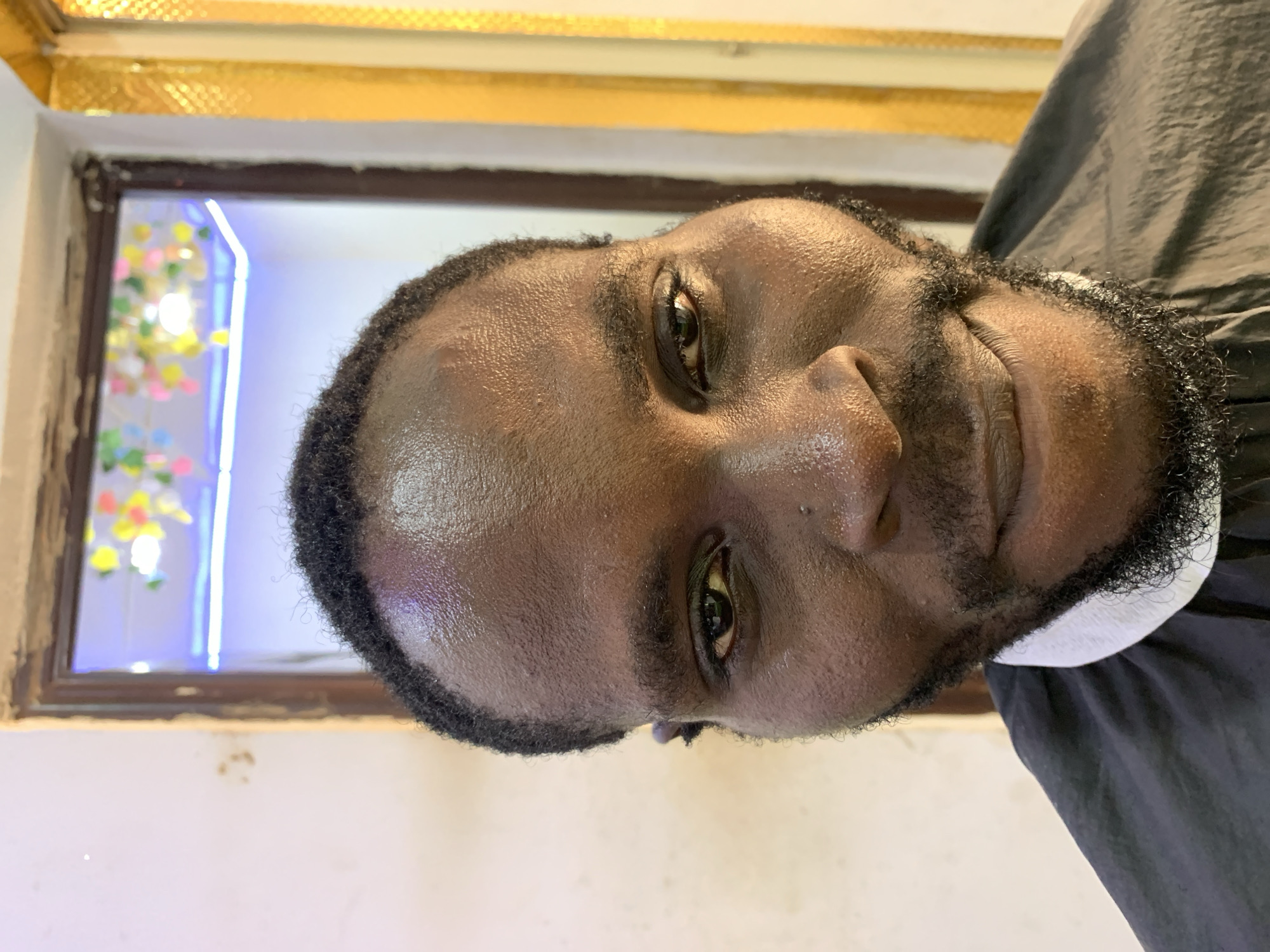 A medium close-up shot depicts a Black man with a closely cropped afro, a beard, and a haircut. The man's skin appears to be slightly shiny. He is wearing a black barber's cape with a white collar. The background features a window frame, with decorative elements and a light source visible through the window. The setting appears to be indoors. The image's location in Ikorodu, Nigeria is not discernible from the visual information provided.