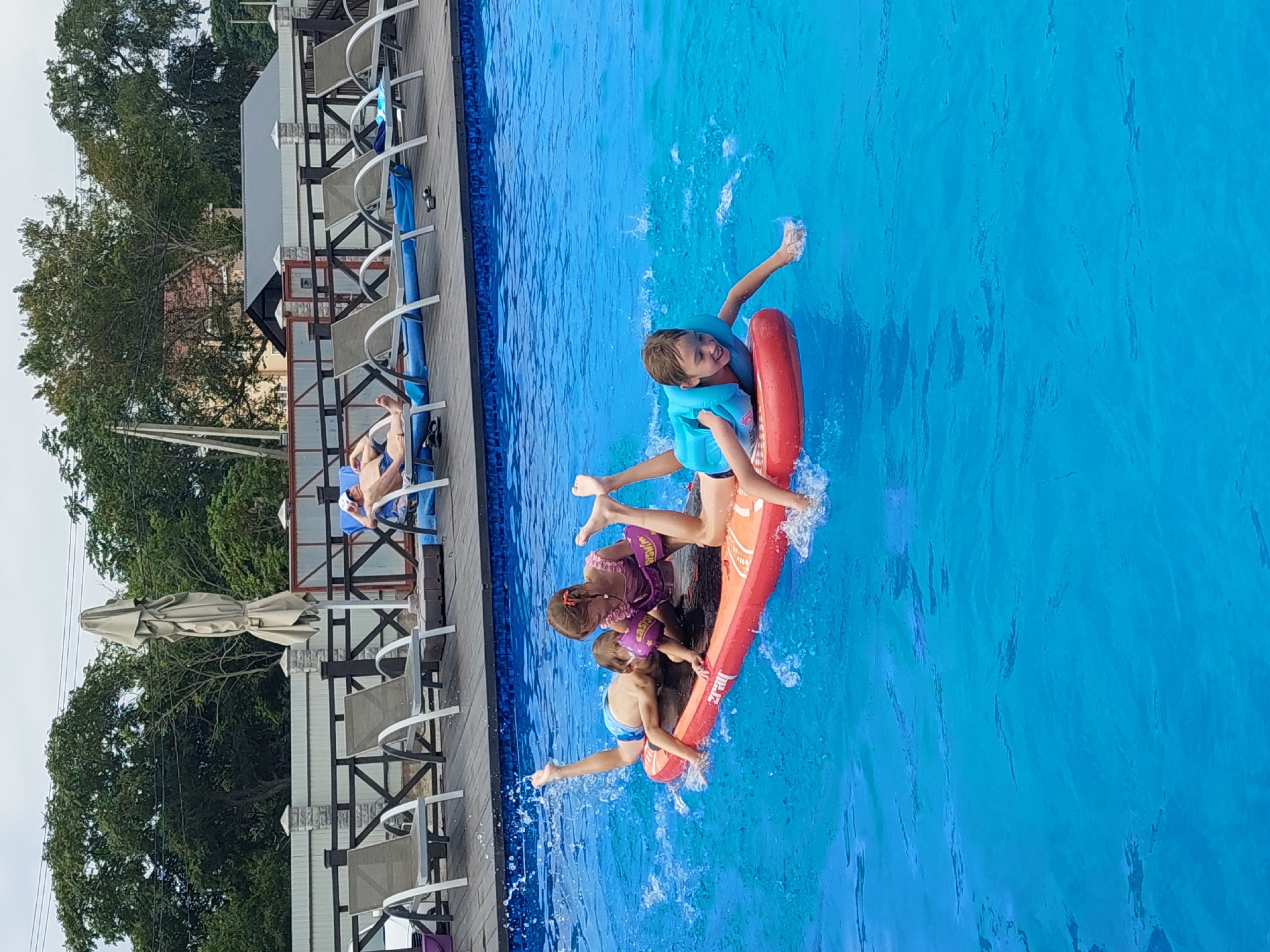 The image depicts a bright, sunny day at an outdoor swimming pool, likely in Andreevka, Russia. The pool water is a vibrant blue, with visible ripples and splashes indicating activity. In the foreground, three children are engaged in play on a large, orange inflatable raft. One child, wearing a blue life vest, is positioned at the front of the raft, paddling with their hands and smiling. Another child, in a pink swimsuit and purple arm floaties, is sitting upright on the raft, facing towards the camera. A third child, wearing blue swim trunks, is lying on the raft, with their legs kicking up water. The raft is positioned in the middle of the pool. In the background, a wooden deck area surrounds the pool, furnished with lounge chairs. Some of these chairs are occupied by adults who are relaxing. Further in the background, trees and buildings are visible, suggesting the pool is situated in a residential or recreational area. An umbrella is folded and standing on the deck. The overall atmosphere is one of leisure and fun.