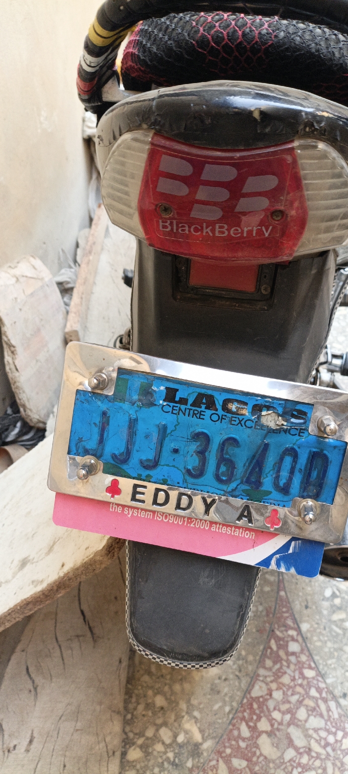 The image captures a close-up view of the rear section of a motorcycle or scooter, possibly parked in an outdoor setting in Kaura Goje, Nigeria. The primary subject is the vehicle's tail light assembly and license plate.
The tail light features a red lens with a prominent "BlackBerry" logo, consisting of the triple B symbol and the word "BlackBerry" beneath it. Below the tail light, a blue license plate is attached within a metallic frame. Visible text on the plate includes "LAGOS" (partially obscured) and "CENTRE OF EXCELLENCE" at the top, and the alphanumeric sequence "JJJ-36400D" centrally. Below the license plate, a pink and blue card or sticker is partially visible, bearing the text "EDDY A" next to a red clover-like symbol, followed by "the system ISO9001:2000 attestation".
The motorcycle's rear fender is black plastic, and parts of the seat or luggage rack are visible above, covered with black netting that has a red mesh pattern. The background on the left shows a rough, light-colored wall or concrete structure, with some debris on the ground. The ground beneath the vehicle is a tiled surface with a mosaic pattern of reddish and beige tones. No activity is taking place, and the scene appears static, suggesting the vehicle is stationary. The wear and tear on the vehicle parts, particularly the faded license plate and scratches on the fender, indicate regular use.