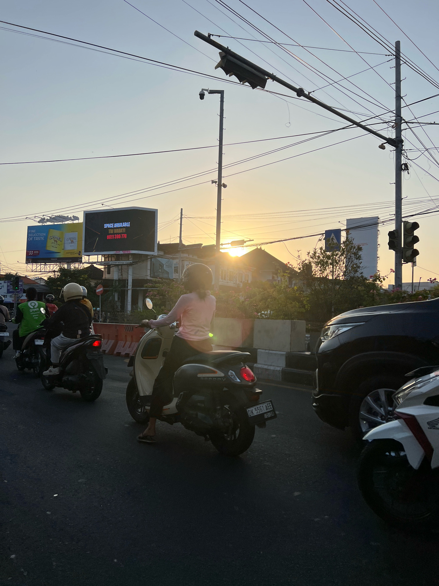 The image depicts a street scene in Kuta, Indonesia, during sunset. The sky is a clear, pale blue with the sun peeking through buildings on the right, casting a warm glow. Multiple motorcycles are present, suggesting traffic congestion.
In the foreground, a woman in a pink shirt rides a white scooter, her back to the camera, wearing a helmet. To her left, a black SUV is partially visible. Behind her, another person on a black scooter also rides forward. Further back, a person in a green "gojek" uniform is on a black scooter, and another individual on a black scooter with a white helmet is in front of them.
A large digital billboard is visible in the background, displaying advertisements. One side shows yellow packaging for Camel cigarettes with the text "FINE SELECTION OF TASTE" and a price of "33.000". The other side of the billboard shows "SPACE AVAILABLE" and a phone number, "0811 396 778".
The street is paved, and on the right side, concrete barriers with black and white stripes are present. Traffic lights are visible overhead, along with a network of power lines and streetlights. The overall impression is a busy urban environment during the late afternoon or early evening.
