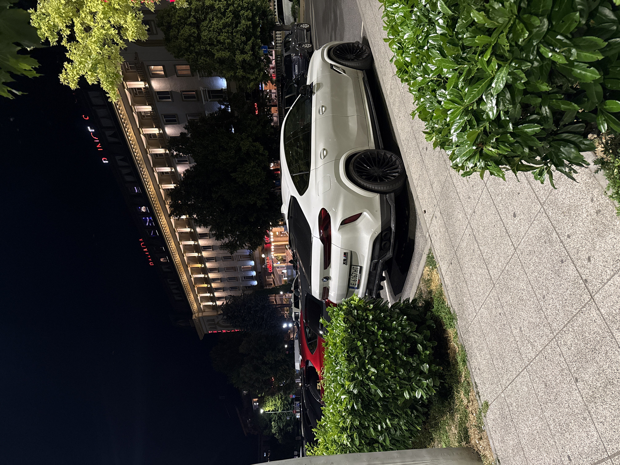 A white BMW SUV is parked on a sidewalk next to a building at night. The car has black rims and tinted windows. Red neon lights spell out "CANADA" on the building. Trees are visible in front of the building, and green bushes line the sidewalk. The scene appears to be in an urban area. The license plate of the car is visible and reads "E 6167 HT". The time of day is night, and the weather appears to be clear.
