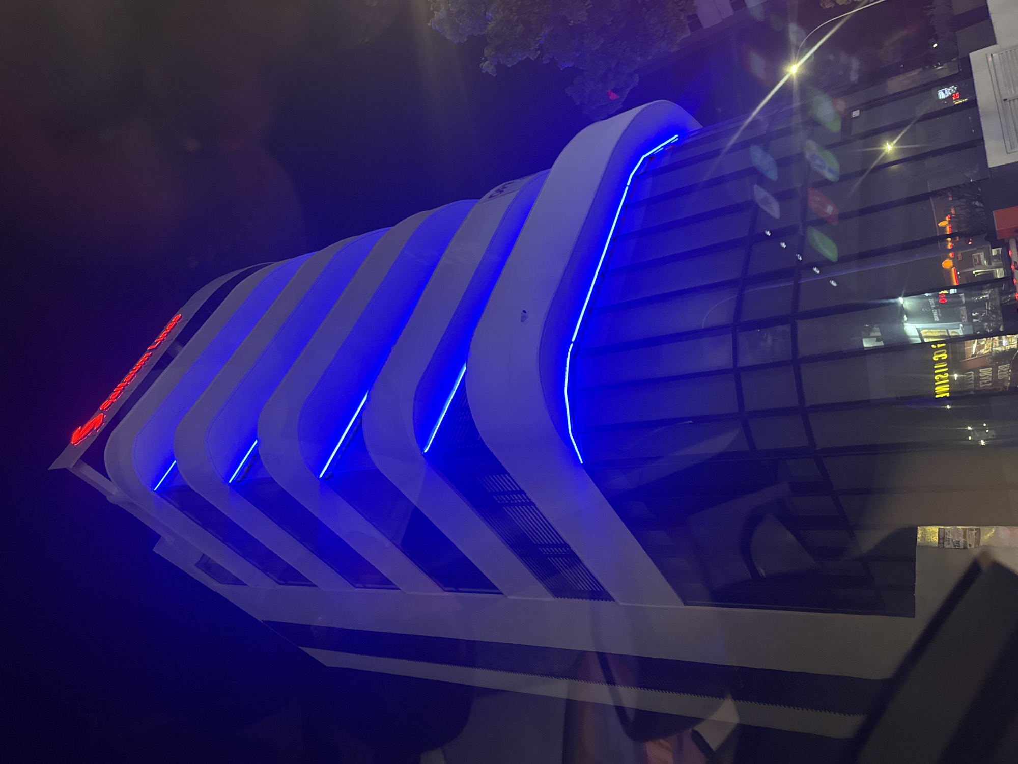 The image shows a modern, multi-story building at night in Limassol, Cyprus. The building has a white facade with curved horizontal elements illuminated by bright blue LED lights. The top of the building features red neon lettering that spells out "Sunfresh". The lower portion of the building is covered in dark, reflective glass, which shows reflections of streetlights and possibly businesses across the street. A streetlamp with a bright yellow glow is visible on the right side of the image, and a tree with dark foliage is partially visible behind the building. The overall impression is a contemporary urban scene with striking architectural lighting.