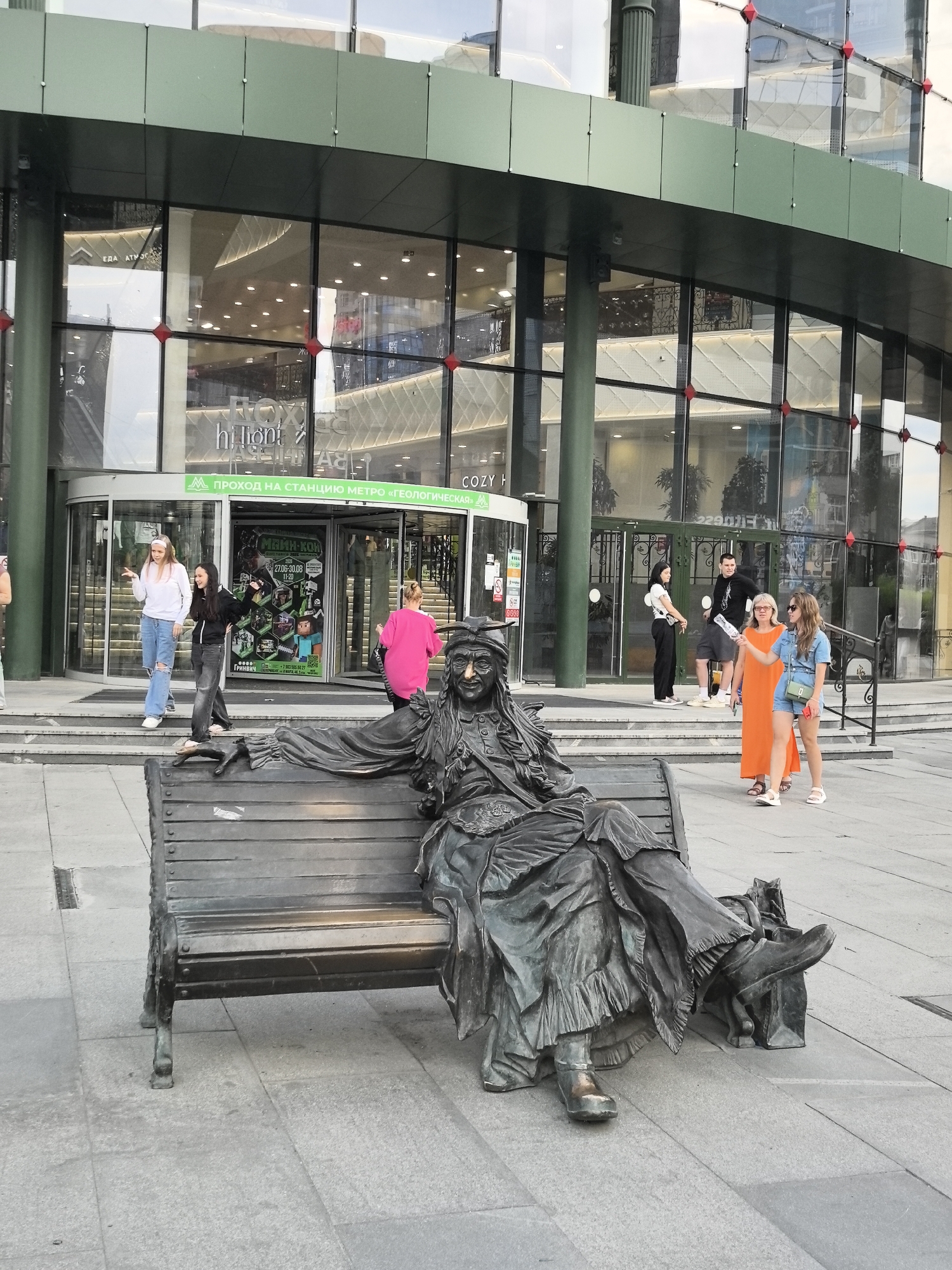 A bronze sculpture of an elderly woman wearing a hat, long dress, and boots is seated on a park bench. She is positioned in the foreground of the image, facing slightly to the left. Behind her, a modern building with large glass windows and green accents serves as a backdrop. The building appears to be a shopping center, with an entrance featuring a revolving door and signage that includes "HILONT" and "COZY H". People are visible in the background, entering and exiting the building and walking on the paved area. One group of people includes two young women, one in ripped jeans and a white top, the other in dark clothing. Another group consists of a man in a black t-shirt and shorts, a woman in a long orange dress, and a young woman in a denim outfit. The overall scene suggests an urban environment in Yekaterinburg, Russia, specifically outside a commercial establishment.