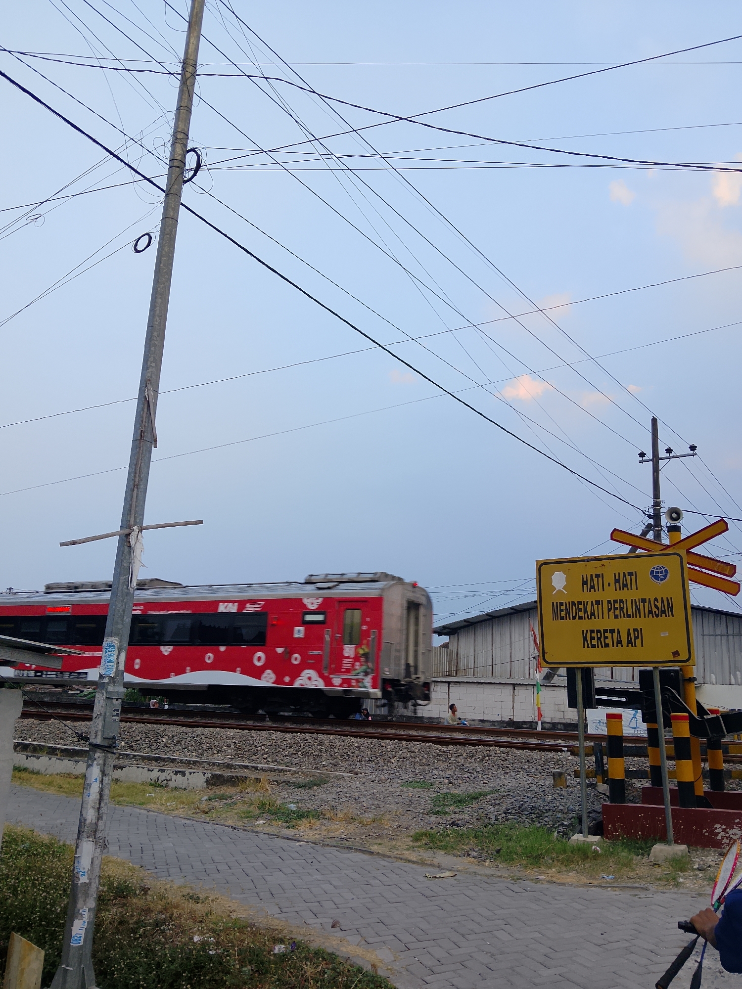 An eye-level, wide shot captures a railroad crossing in the city of Ngadrin, Indonesia. The sky is a pale blue with a few wispy clouds. A red train with white circular patterns is in motion, blurred due to its speed as it passes through the crossing. To the right, a yellow sign with black lettering warns of an approaching railway crossing. Red and yellow striped barriers are lowered, indicating the train's approach. The foreground shows a paved walkway and some gravel next to the railroad tracks. A metal utility pole stands on the left side of the frame, covered in various stickers and sporting a horizontal wooden support. In the background, industrial-style buildings line the far side of the tracks, and a few individuals are visible near the train. The overall atmosphere is that of an ordinary day at a functional, active railway intersection.