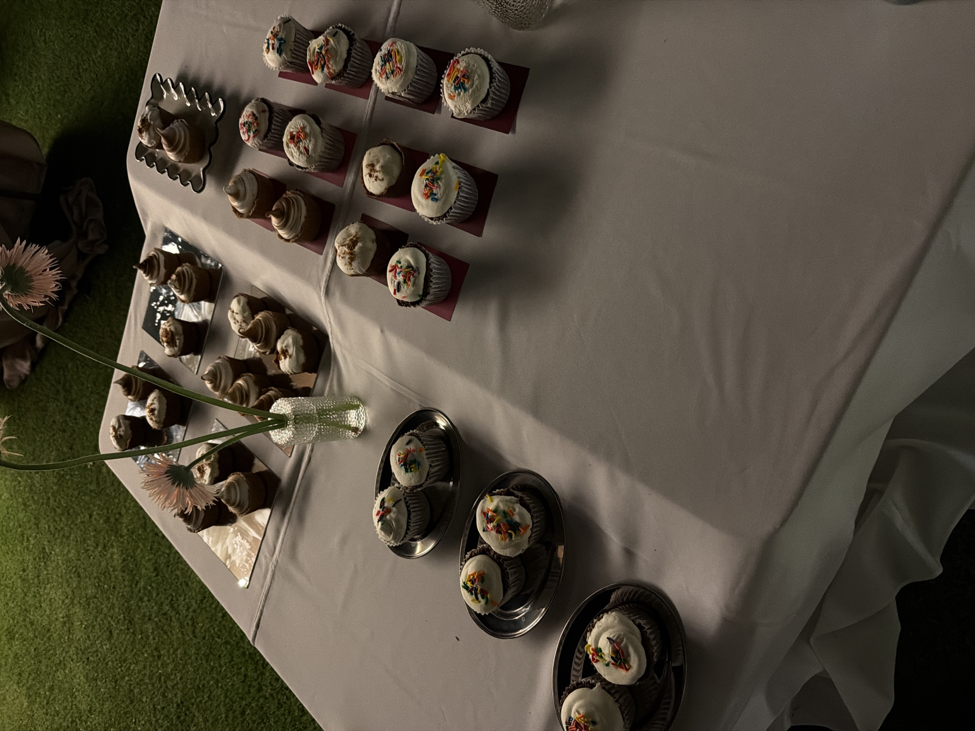 The image shows a table laden with desserts, likely at an event. The table is covered with a white cloth. A selection of cupcakes with white frosting and colorful sprinkles are arranged in rows, some on individual squares, others on small platters. In the foreground, on the left, are small pastries arranged on rectangular mirror tiles, near a vase containing flowers. The background shows a patch of artificial green turf. The location is presumed to be San Bernardino Tlaxcalancingo, Mexico. No people are visible. The focus is on the display of desserts.