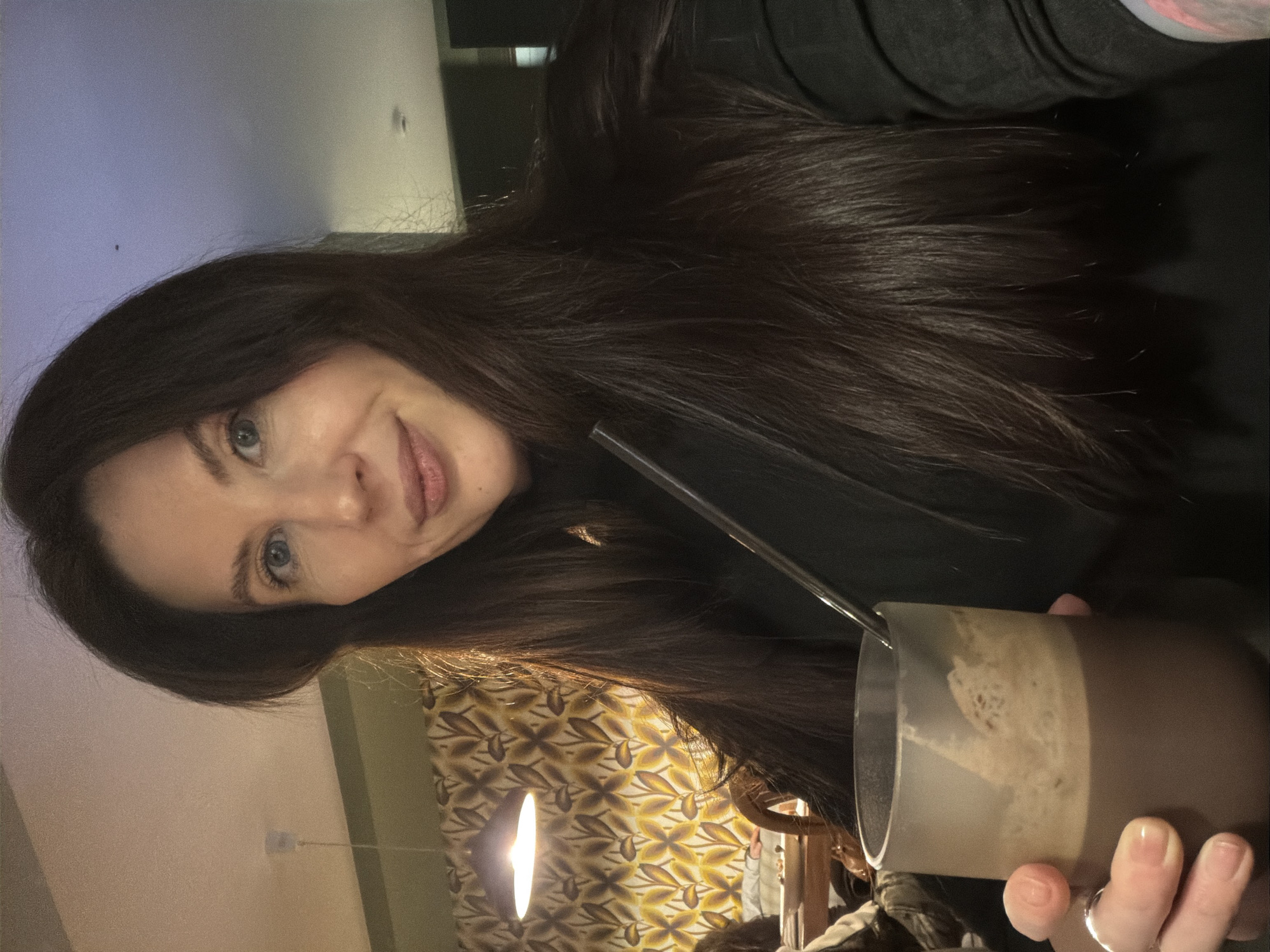 A person with long, dark hair is holding a brown and clear glass of what appears to be a foamy beverage with a straw in it. The person has light skin and blue eyes and is wearing a dark-colored top. The background includes a patterned wallpaper in shades of brown and yellow, a light fixture, and a glimpse of a neutral-colored wall. There is no information to confirm it is taken in Perm, Russia.