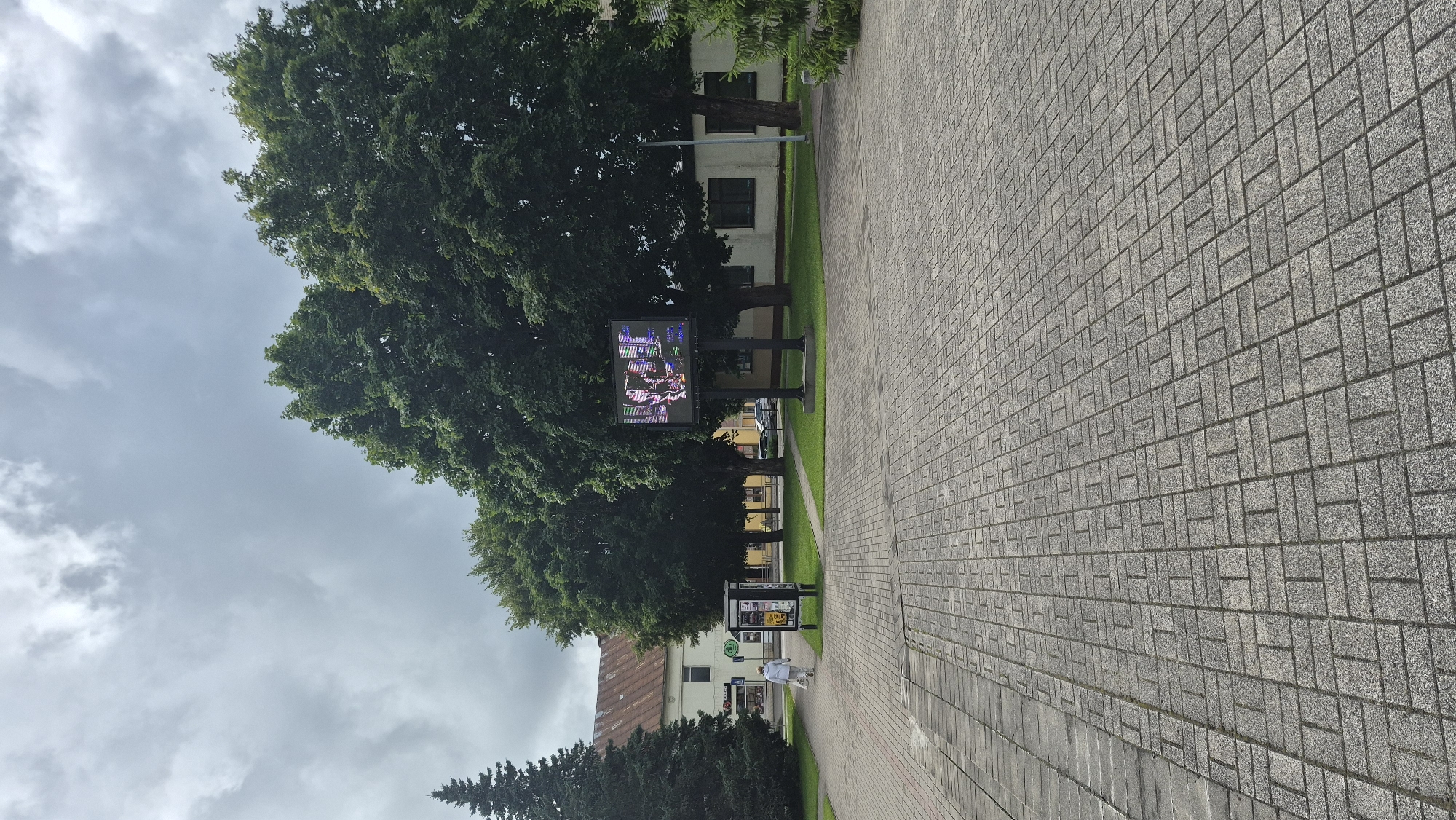 The image captures an outdoor scene in Krāslava, Latvia, likely a public square or plaza. The ground is paved with grey rectangular stones laid in a basketweave pattern. A person, dressed in a light-colored outfit and carrying a white bag, walks away from the camera towards a building on the left side of the frame. This building appears to be a commercial establishment with signage visible, including a sign with red lettering. Adjacent to the building is a tall evergreen tree.
Dominating the mid-ground are several large, mature deciduous trees with dense green foliage. In front of these trees, a large digital billboard displays a colorful image. The billboard is supported by two dark poles. Behind the trees and billboard, a low-rise building with a light-colored facade and darker windows is visible. The sky above is overcast with grey clouds. To the far right of the frame, part of another evergreen tree is visible.