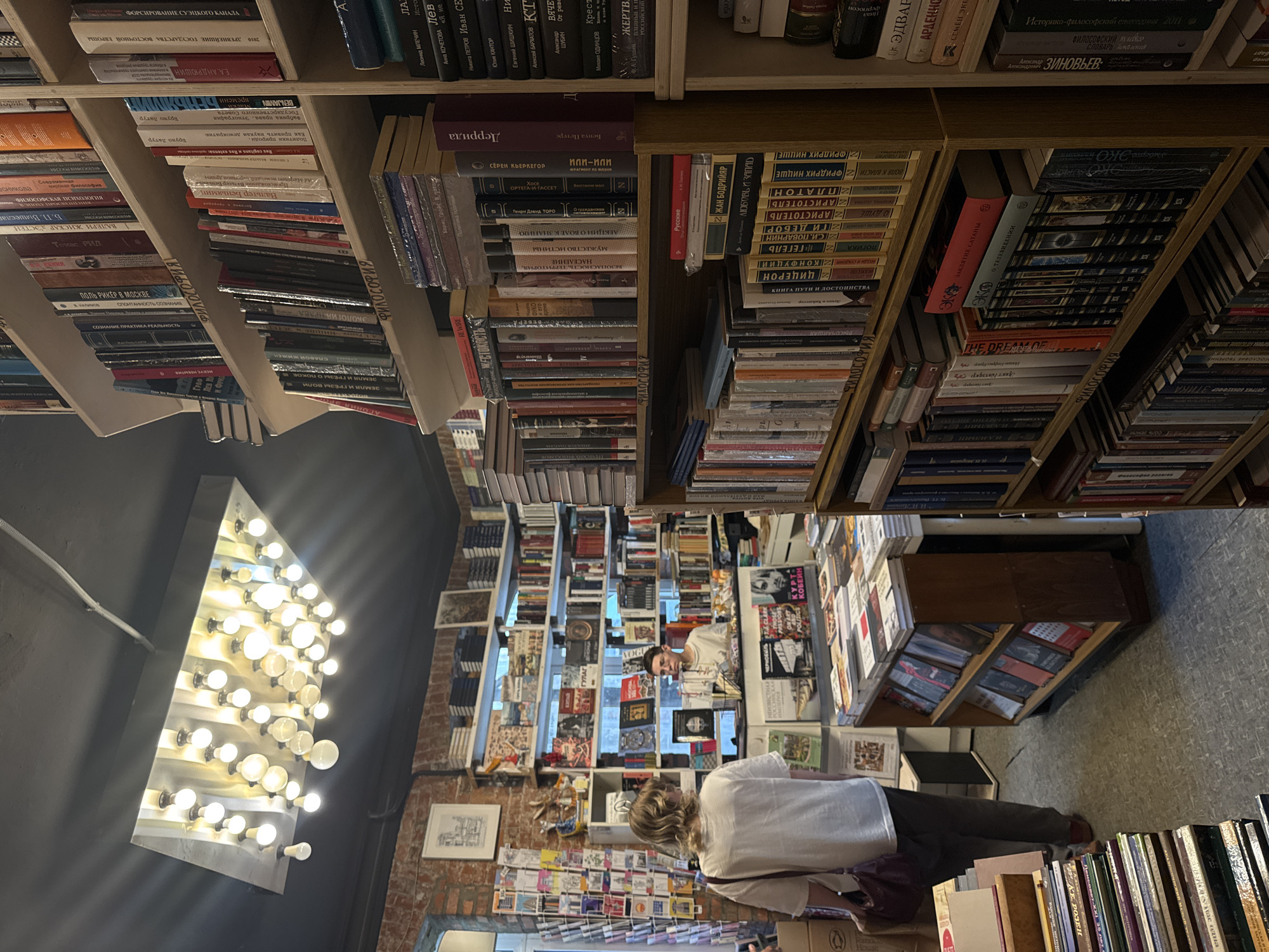 This image depicts the interior of the "Фаланстер" (Falanster) bookstore, located at Maly Gnezdikovsky Lane, 12, Moscow, Russia.
The scene is an indoor, well-lit view of a densely packed bookstore. The dominant feature is a series of tall, wooden bookshelves lining the walls and forming aisles, overflowing with books predominantly in Russian. A unique, large, rectangular overhead light fixture, constructed from a metallic panel and fitted with approximately 30-40 exposed spherical incandescent bulbs, illuminates the central area. The ceiling is dark, contrasting with the light fixture. The left wall features exposed red brick, extending towards the back.
Two individuals are present. One person, with light-colored hair, wearing a white t-shirt and dark pants, and carrying a dark shoulder bag, stands facing away from the camera, looking towards a checkout counter in the mid-ground. Her back is mostly visible. The second person, wearing glasses and a light-colored top, is positioned behind a wooden checkout counter, facing the first individual.
Various books are stacked on shelves, tables, and floor displays. Visible Russian text on book spines includes "ФИЛОСОФИЯ" (PHILOSOPHY) on multiple shelves, and author names such as "ГЕГЕЛЬ" (Hegel), "НИЦШЕ" (Nietzsche), "ПЛАТОН" (Plato), "ЖАН БОДРИЙАР" (Jean Baudrillard), and "КУРТ КОБЕЙН" (Kurt Cobain) are discernible on various titles. A rack displaying postcards or similar small items is partially visible on the far left against the brick wall. The floor is covered with gray, textured tiles.