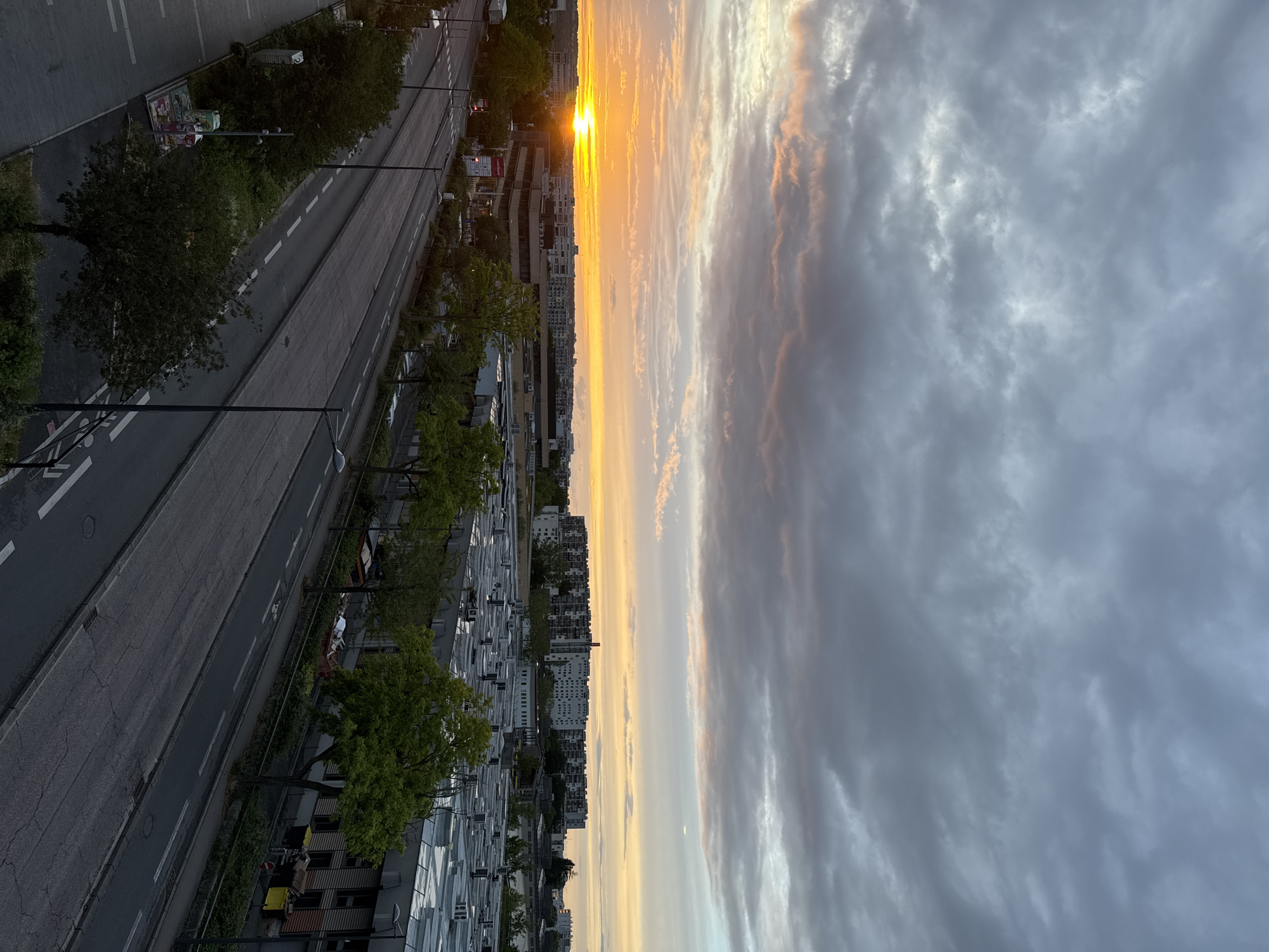 This is a photograph of a street and surrounding urban landscape in Fresnes, France, at sunset. The sky is filled with dramatic clouds, tinged with orange and yellow hues from the setting sun, which is visible on the left side of the frame, low on the horizon. The city features a mix of buildings, including apartment complexes and industrial or commercial structures with flat roofs. Trees line the street, their leaves green and full. The street itself is paved and appears to have multiple lanes, with visible lane markings. There are streetlights and traffic signals visible. No people are clearly discernible in the image. The overall impression is of a quiet urban scene during twilight.
