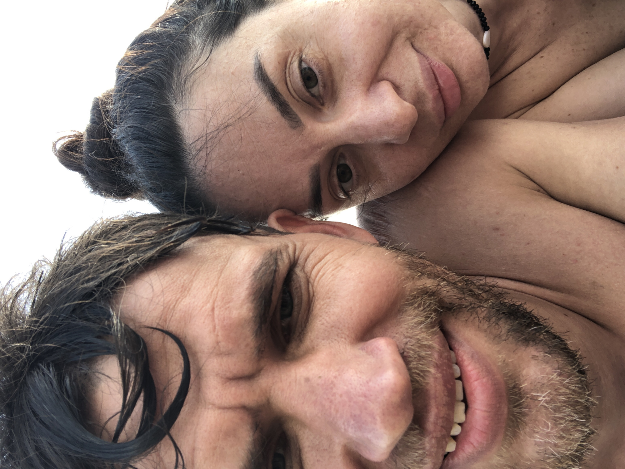This image, captured in the city of Sestroretsk, Russia, presents a close-up, selfie-style portrait of two individuals, a male and a female, positioned in close proximity. The lighting is bright and even, suggesting an outdoor setting or diffused indoor light with a plain, light background.
The male, occupying the left side and foreground, is partially cropped from the top of his head to below his chin. He has dark, wet, disheveled hair, a light brown beard, and a visible mustache. His eyes, partially obscured by shadows, look slightly upward, and he displays a subtle, open-mouthed smile, revealing a slight gap between his upper front teeth. Wrinkles are evident around his eyes and across his forehead. His skin appears tanned with visible pores and slight redness on his nose.
The female is situated to the right and slightly behind the male, leaning into his shoulder. Her dark hair is gathered into a high bun, with some loose strands visible around her face. She has defined eyebrows, dark eyes that look directly at the viewer, and a gentle, closed-mouth smile. Her skin also appears tanned, with visible pores and some freckling. She wears a dark beaded necklace with a central white element. Both individuals appear to be topless or wearing minimal swimwear, exposing bare shoulders and upper chests, indicating a casual, warm environment.