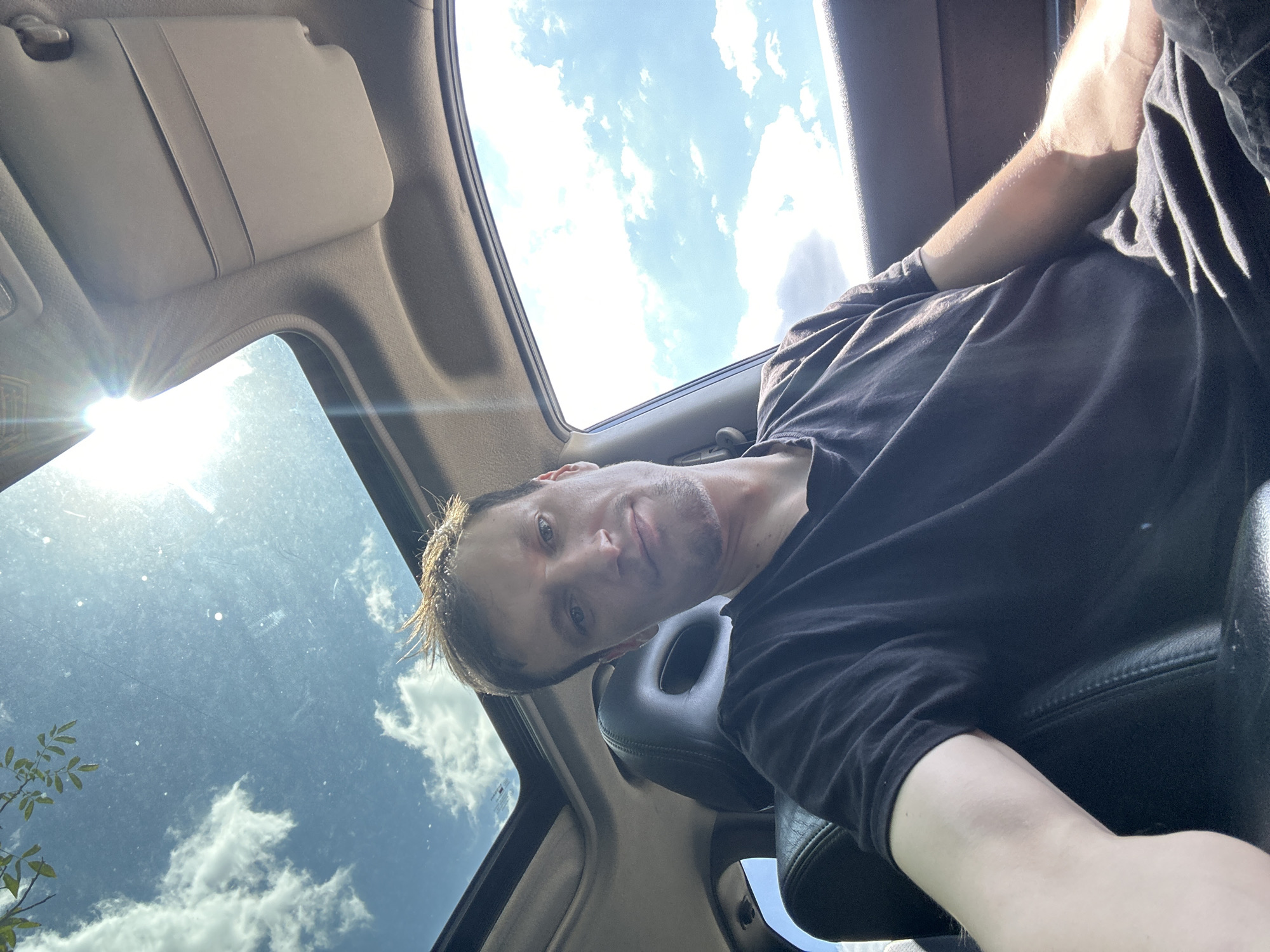 A man with light brown hair, wearing a black t-shirt, is sitting in the driver's seat of a car. Sunlight streams through the open sunroof, casting a bright glare. The sky outside is blue with scattered white clouds, and a tree branch with green leaves is visible against the sky. The interior of the car includes a black leather seat and the car's visor. The scene appears to be captured from a selfie perspective.
