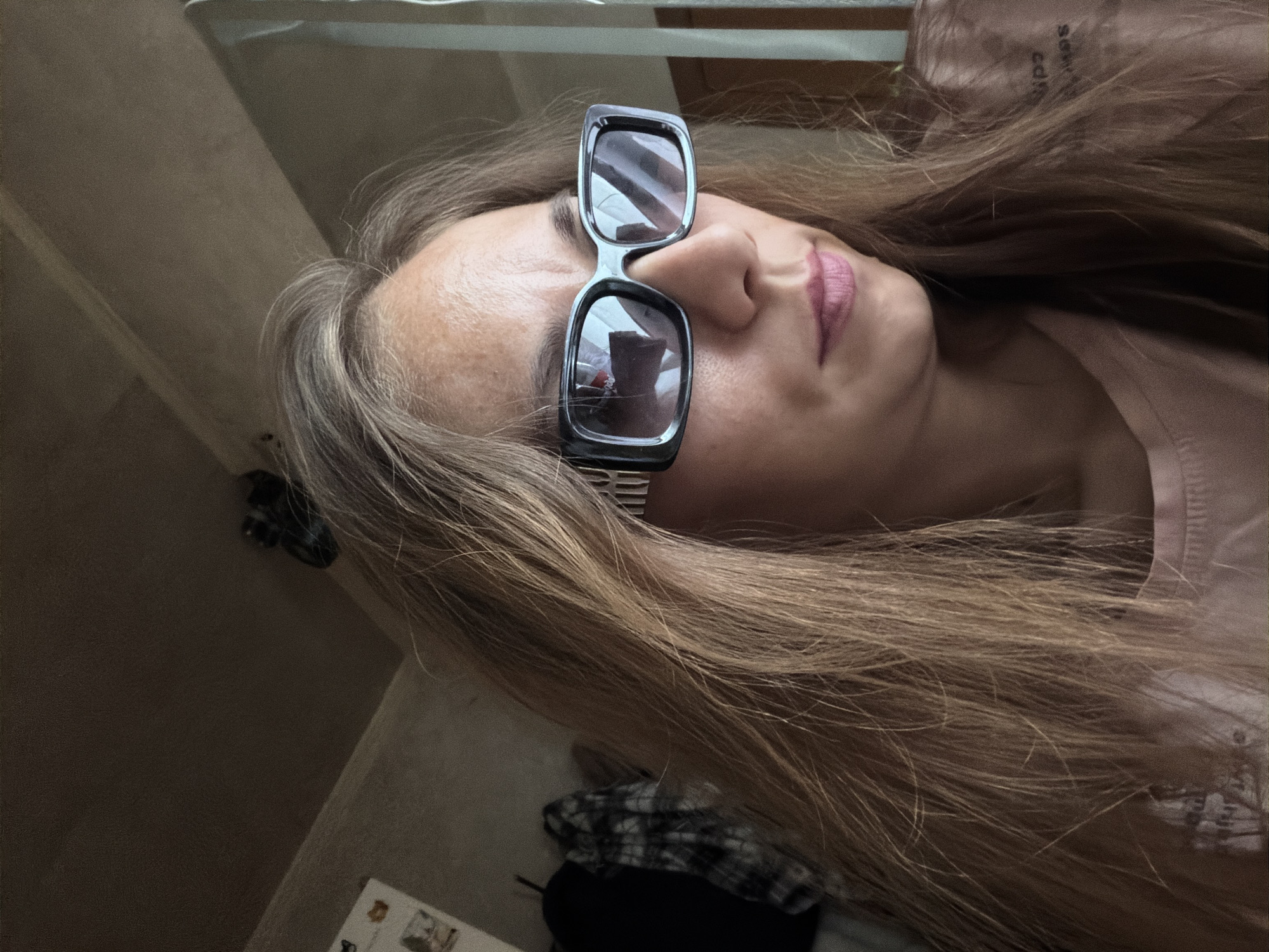 This is a close-up, medium shot photograph of a woman wearing sunglasses indoors. The woman has long, brown hair that is parted in the middle and falls around her face. She is wearing a pink t-shirt. The sunglasses are black and rectangular with thick frames. The background of the image is dimly lit and appears to be a room with textured walls and a ceiling. On the left side of the background, there is a glimpse of a black garment hanging on a hook and a black and white plaid shirt. There is also a small picture frame on the wall. The overall impression is that this is a casual, indoor portrait. There is no indication of the city of Tambov, Russia, or any specific location within it.