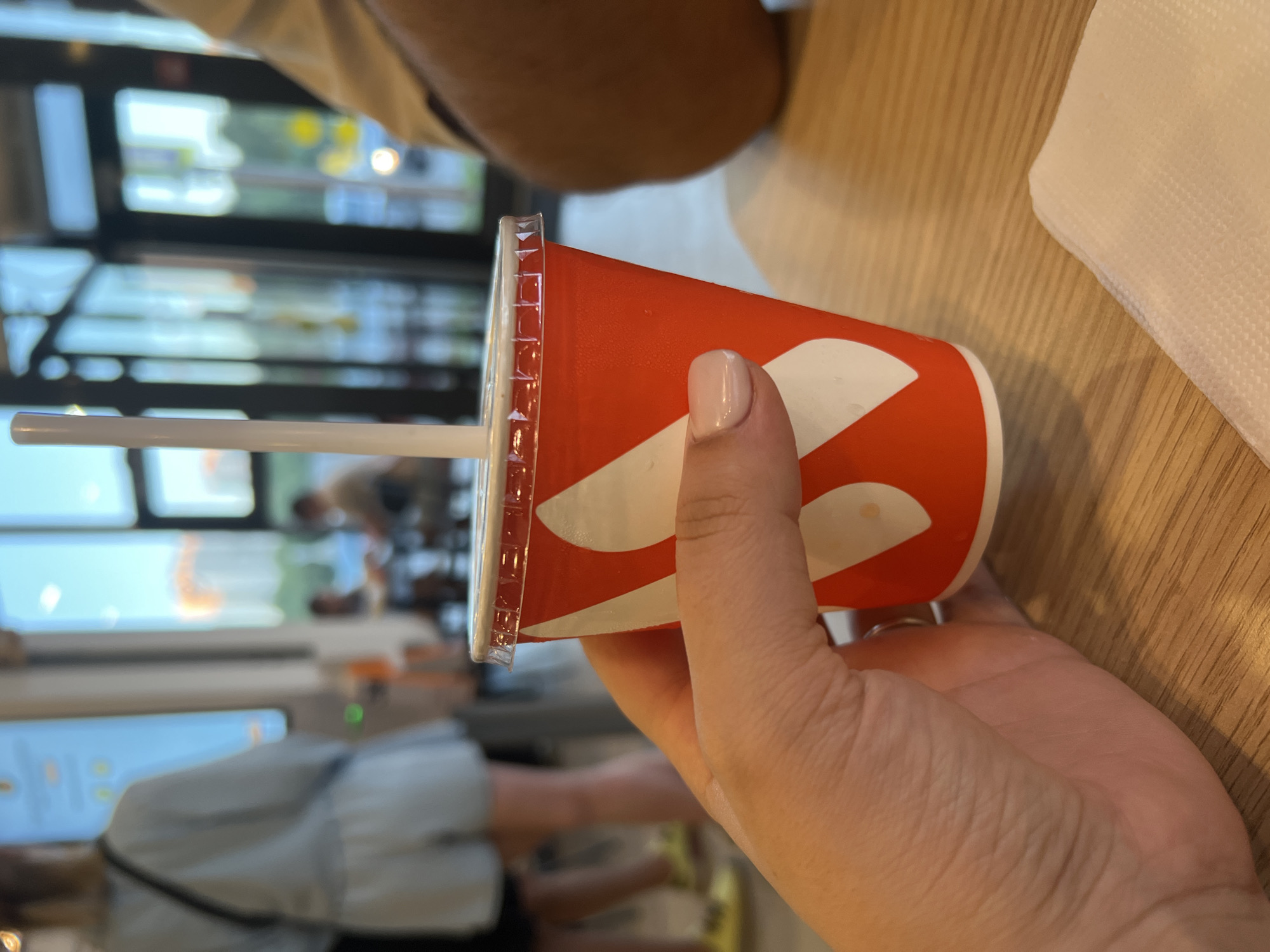 A woman's hand holds a red paper cup with a white swirl design and a clear plastic lid. A white straw extends from the cup. The cup is on a wooden table, next to a white napkin. In the blurred background, people are visible inside a modern establishment with large windows, suggesting a fast-food restaurant or cafe. The lighting indicates it is daytime, and the presence of buildings in the distant background hints at an urban environment, potentially in Yekaterinburg, Russia.