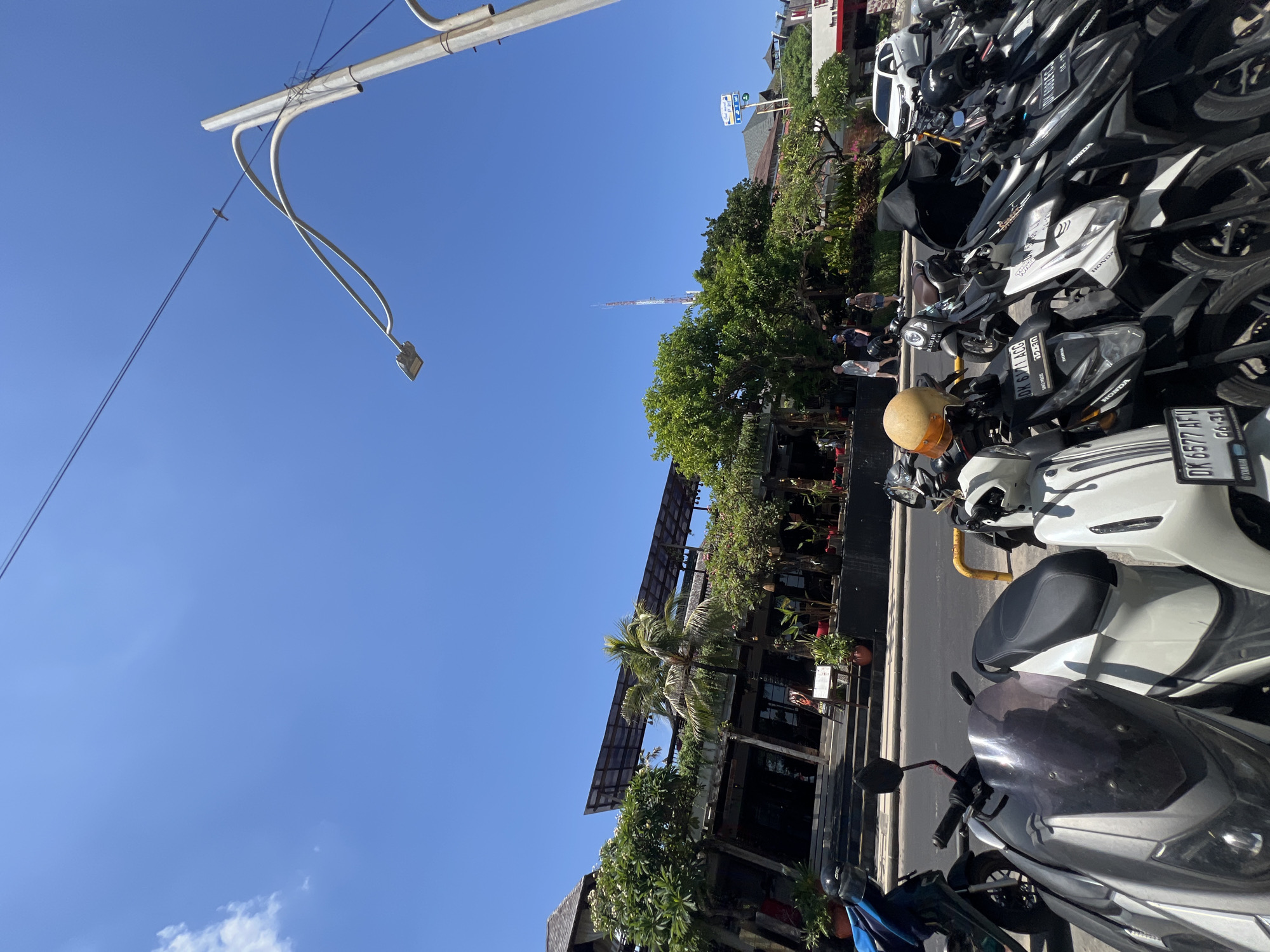 In Kuta, Indonesia, under a clear blue sky, a row of parked scooters occupies the foreground on a paved street. A white car is visible in the background. Behind the scooters, a multi-story building with a dark roof structure and an open-air design is lined with trees and potted plants. Several individuals are walking near the building's entrance, and a tall, white and red tower stands in the distance. A street lamp with a modern design is also present, with power lines stretching across the sky.