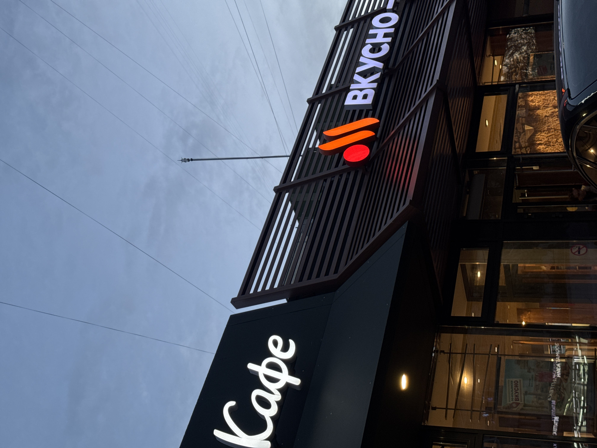 This media file depicts the exterior of a cafe, likely in Saint Petersburg, Russia, under a cloudy, overcast sky. The building features a modern architectural design with dark wood-slatted cladding and large glass windows. Prominently displayed are illuminated signs. One sign, in white Cyrillic script, reads "ВКУСНО," meaning "tasty" or "delicious." Below this, a stylized orange and red logo is visible, possibly representing flames or abstract shapes. Another sign, in a flowing white script, reads "Кафе," meaning "cafe." Through the glass windows, interior lighting and furnishings are partially visible, suggesting a warm and inviting atmosphere within. The overall impression is of a contemporary establishment ready to serve customers. The time of day appears to be late afternoon or early evening, given the low light conditions and the illumination of the signs. The weather seems cool and potentially damp, consistent with a cloudy sky. No people are clearly visible in the scene.