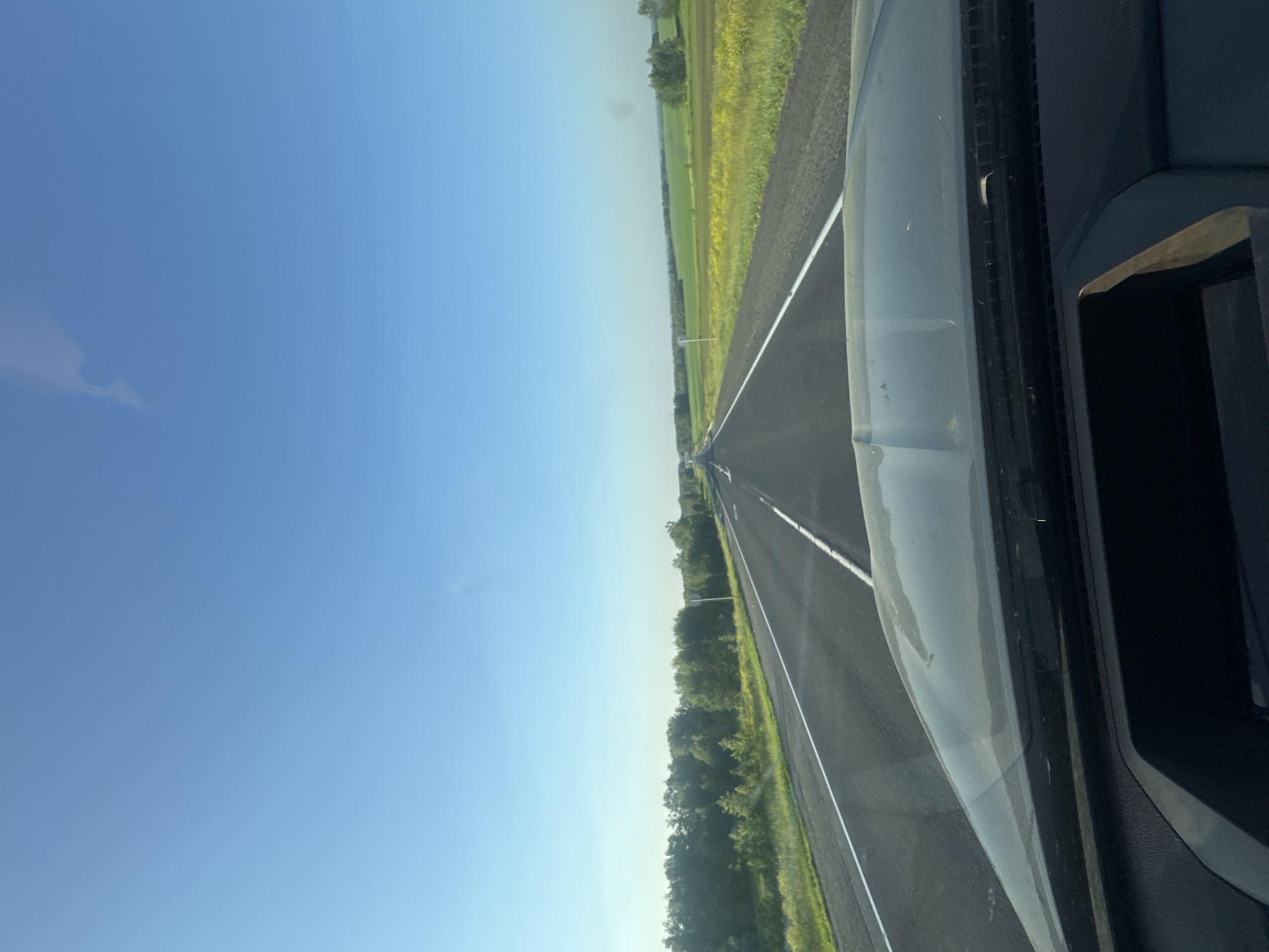 A POV shot from inside a vehicle shows a clear blue sky with a faint wispy cloud. The vehicle is on a paved highway, with white lane markings visible. To the left of the road, there is a line of green trees and foliage, with some yellow wildflowers interspersed. To the right of the road, there are fields of green crops and yellow flowers. The highway stretches into the distance, appearing to lead towards a wooded horizon. The dashboard and hood of the vehicle are visible in the foreground. The scene appears to be a rural landscape in Zeledeevo, Russia, likely during daylight hours given the clear sky.
