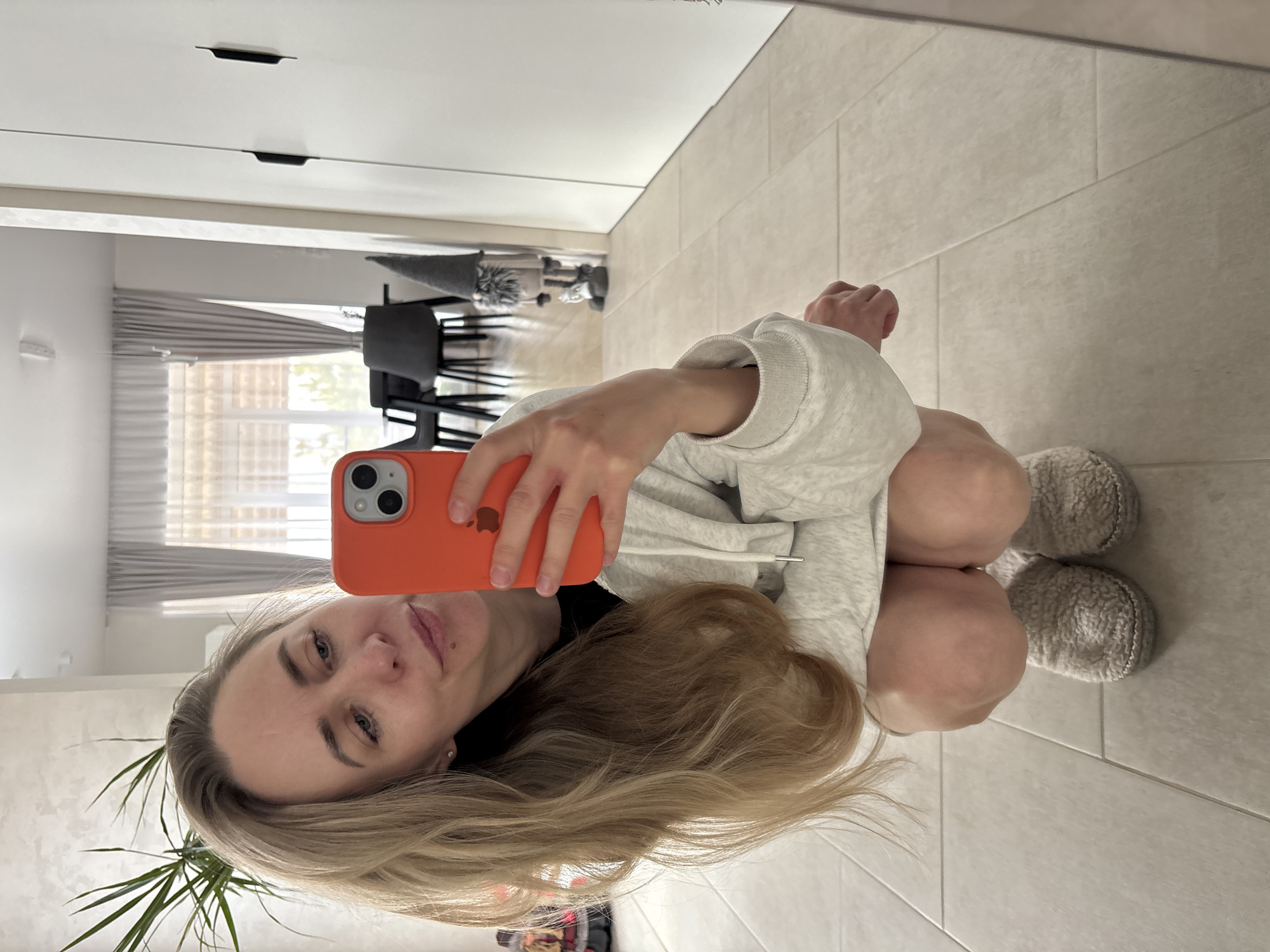 A woman is taking a selfie in what appears to be a mirror reflection. She is wearing a light gray hoodie and fuzzy gray slippers, squatting on a tile floor. She is holding an orange iPhone in her right hand. The reflection shows a glimpse of a room with white cabinets, a black chair, and a window with striped curtains. There is also a potted plant in the lower left corner. The specific location is not possible to determine from this image.