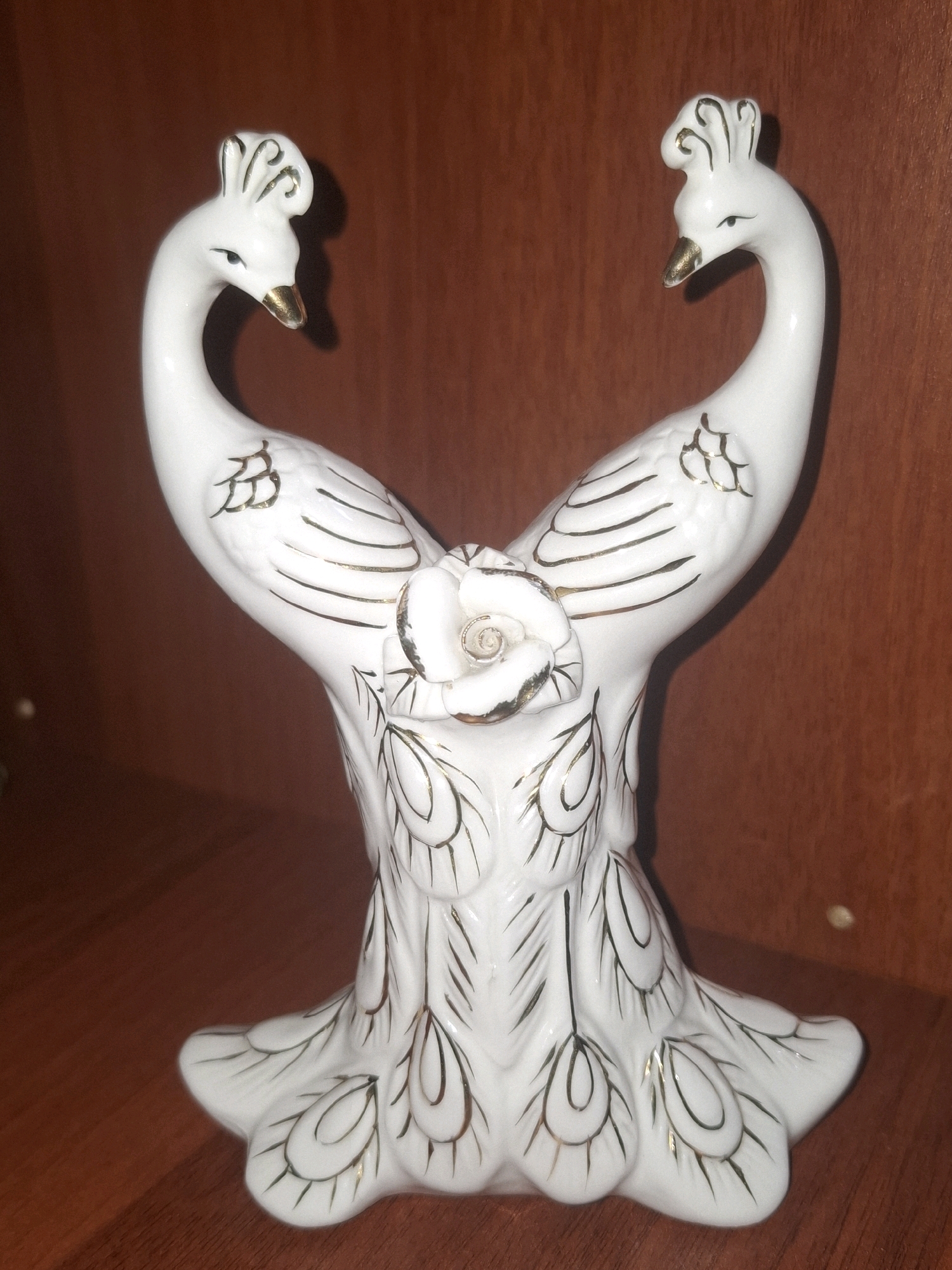 A white ceramic figurine of two peacocks facing away from each other, their necks curved to form a heart shape, stands on a wooden surface. The figurine is adorned with gold trim, highlighting the details of the peacocks' crests, beaks, eyes, wings, and the stylized feathers at the base. A white rose with gold accents is positioned between the two peacocks where their necks meet. The background is a dark brown wooden surface, likely a shelf. The scene is lit from above, casting shadows. The image is a close-up shot of the figurine. There are no people present in the image. This image is not located in the city of Almaty, Kazakhstan.