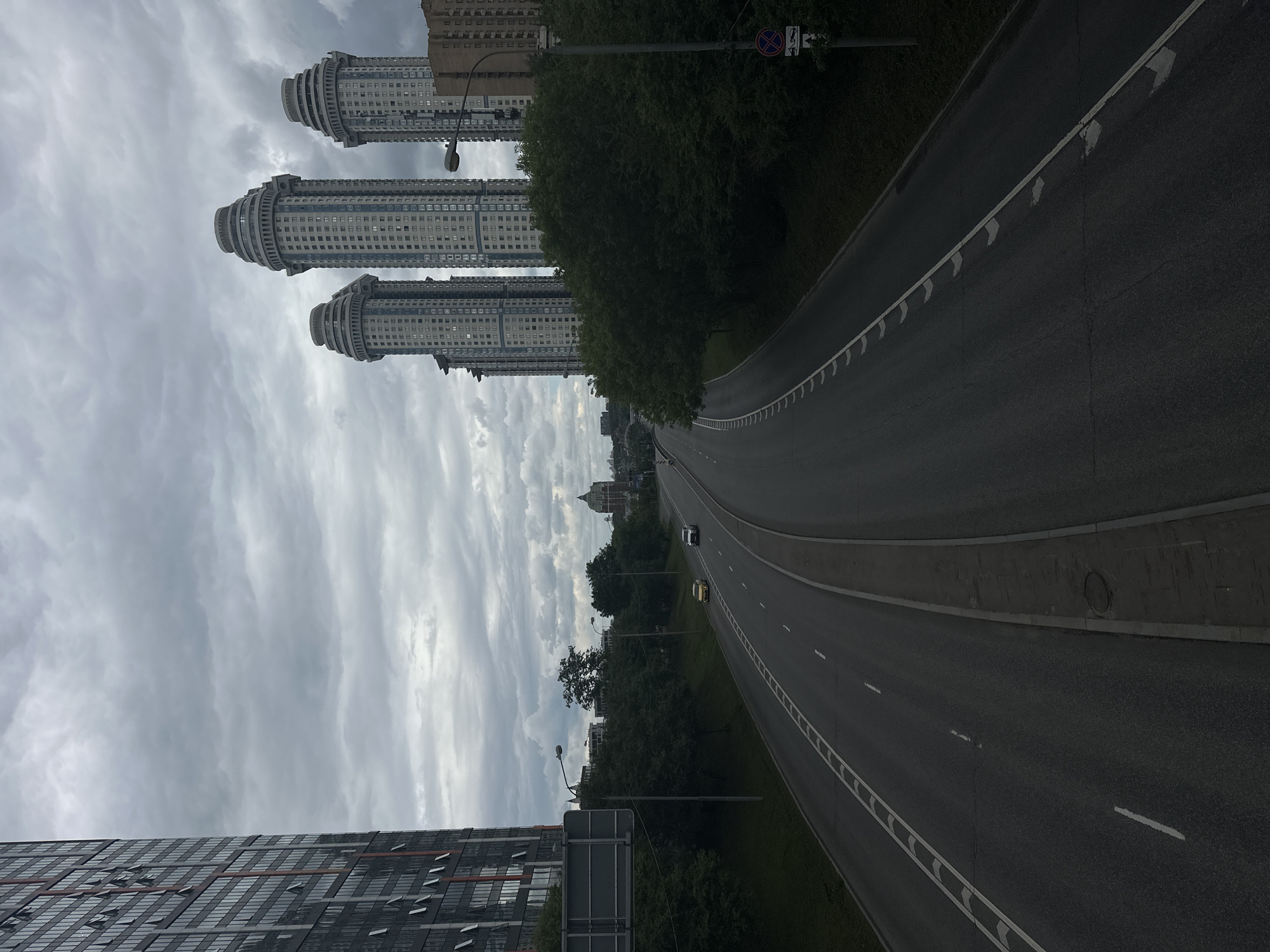 The image depicts a wide, multi-lane asphalt road in Moscow, Russia, with a slightly overcast sky. On the left, a tall, modern skyscraper with numerous windows dominates the frame. In the distance, to the right of the road, a cluster of distinctive cylindrical high-rise buildings with decorative tops are visible, alongside other residential buildings. Green trees line the sides of the road and the areas between the buildings. A single yellow car and a silver car are driving on the road, heading away from the viewer. The scene conveys an urban landscape characterized by contemporary architecture and infrastructure.