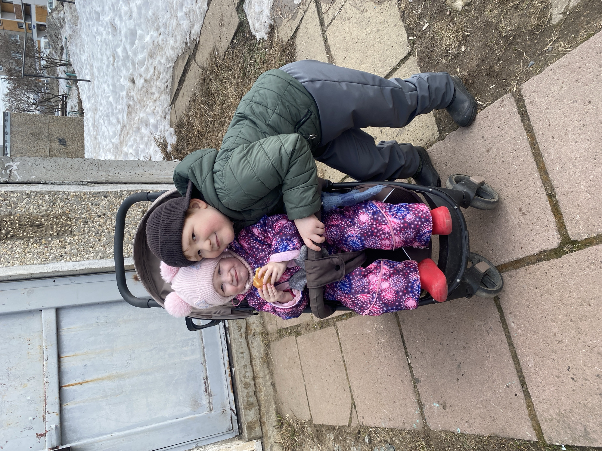 Two children are depicted outdoors on a paved path in Chaikovsky, Russia, during what appears to be late winter or early spring. A younger female child, wearing a pink hat with two pom-poms, a multicolored pink, purple, and blue winter suit with a starburst pattern, and red boots, is seated in a dark-colored stroller, holding a small, round, light-brown bun. She is looking towards the camera with a slight smile. An older male child stands to the right of the stroller, leaning slightly over it with his left arm near the stroller's frame. He wears a dark beanie, a dark green quilted hooded jacket, dark gray winter pants, and black boots, also looking towards the camera with a smile. The immediate ground consists of reddish-brown rectangular concrete paving slabs. To the left, a weathered light gray metal door with rust streaks is set into a concrete wall with exposed aggregate. The background features uneven ground partially covered in melting snow and patches of dry brown grass. Further back, concrete residential buildings, bare deciduous trees, and a distant white vehicle are visible under an overcast sky.
