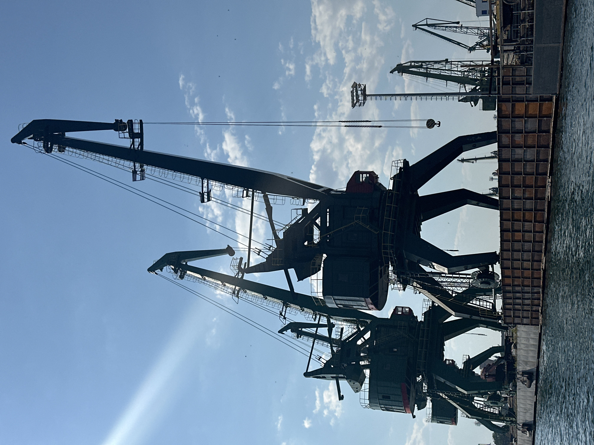 This is a medium shot of a harbor with several large, dark green cranes against a bright blue sky with scattered white clouds and a visible sunbeam. The cranes are silhouetted and appear to be made of metal. The closest cranes are massive, with wide, splayed legs and large, enclosed operator cabins. They are positioned on a dock or barge that is partially submerged in blue water. In the background, there are more cranes of varying sizes and designs, as well as a tall, slender tower with lights at the top. The water in the foreground is choppy and reflects the sky. The scene appears to be industrial, likely a shipping or loading area.