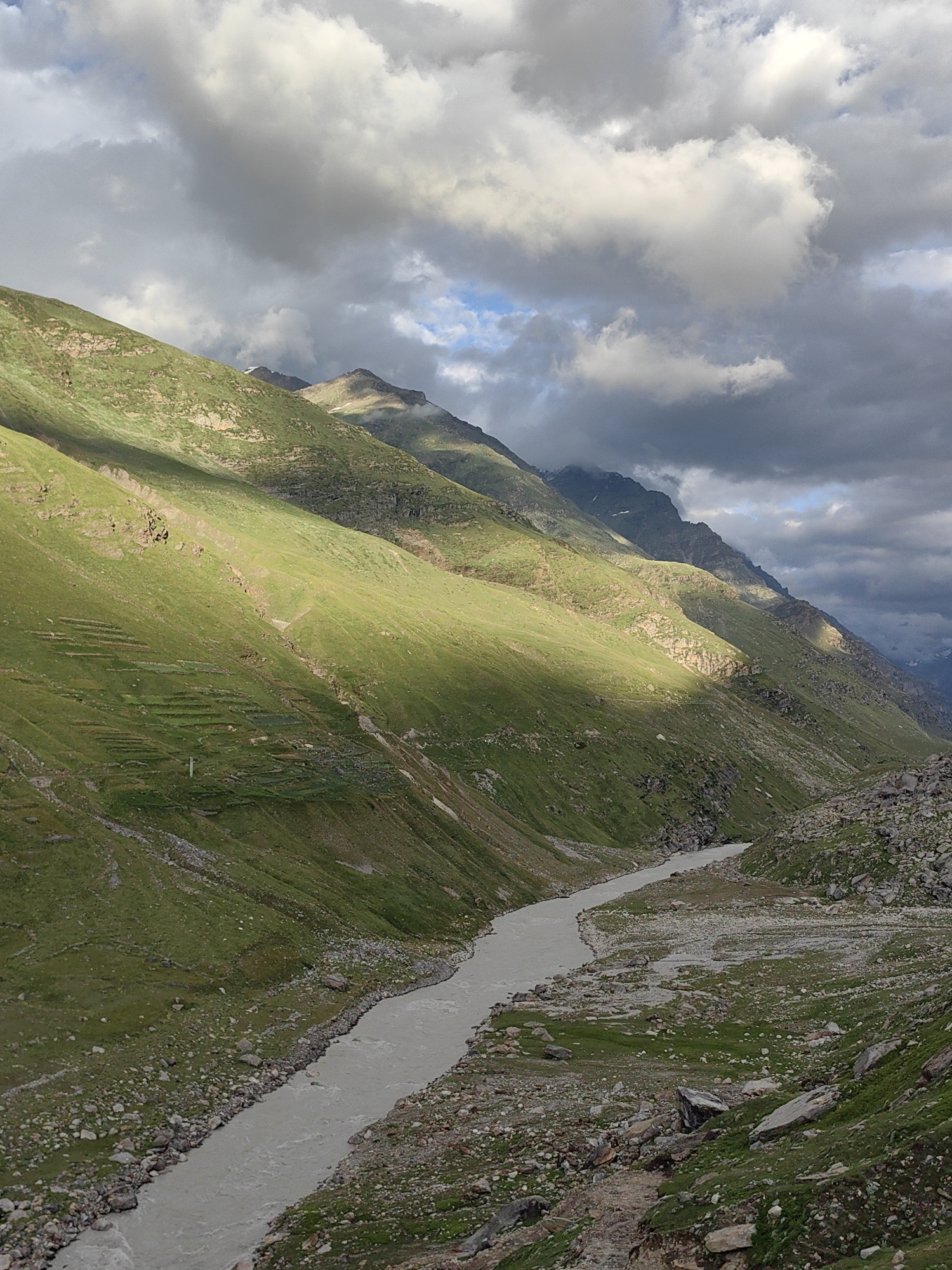The image depicts a dramatic mountain landscape with a river flowing through a valley. The foreground features a wide, silt-laden river snaking through a rocky and grassy riverbed. Steep, verdant mountain slopes rise on either side of the valley, bathed in dramatic shafts of sunlight piercing through a cloudy sky. The slopes are characterized by lush green grass, interspersed with rocky outcrops and evidence of terraced farming or ancient settlements. In the distance, more rugged mountain peaks are visible, some shrouded in clouds. The overall impression is one of a remote, natural environment, possibly the Yari-Khoksar region of India, known for its high-altitude terrain and challenging natural beauty. There are no people or animals visible in the frame.