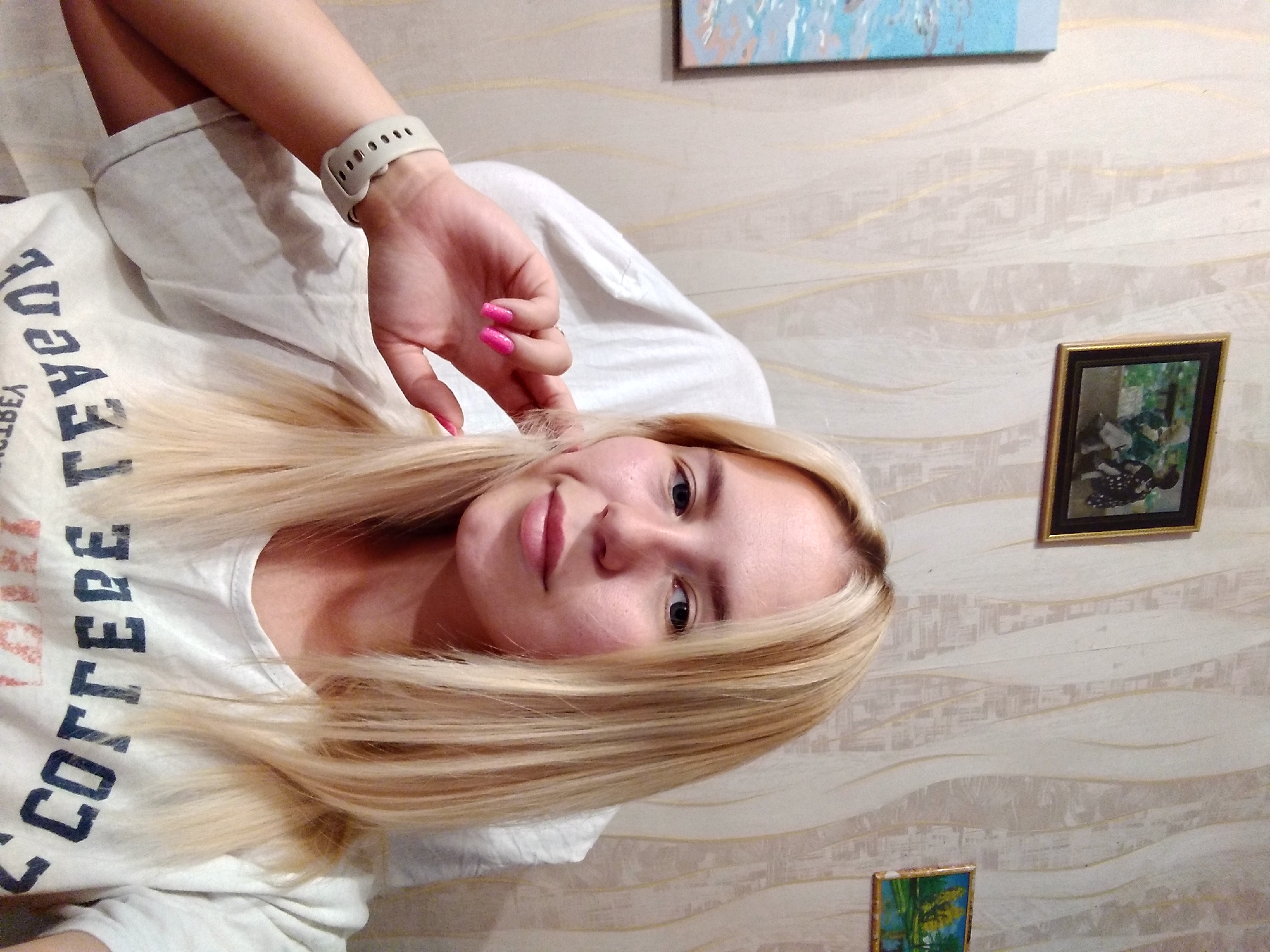 A young woman with blonde hair, wearing a white t-shirt with "COLLEGE" printed on it in large letters, sits in a chair in front of a wallpapered wall. She is wearing a white smartwatch on her left wrist and has bright pink fingernail polish. On the wall behind her are three framed pictures. The one on the far left is a tall, narrow picture with blue and white colors. The middle picture is a landscape scene of trees by a body of water. The picture on the right is a portrait of two people.