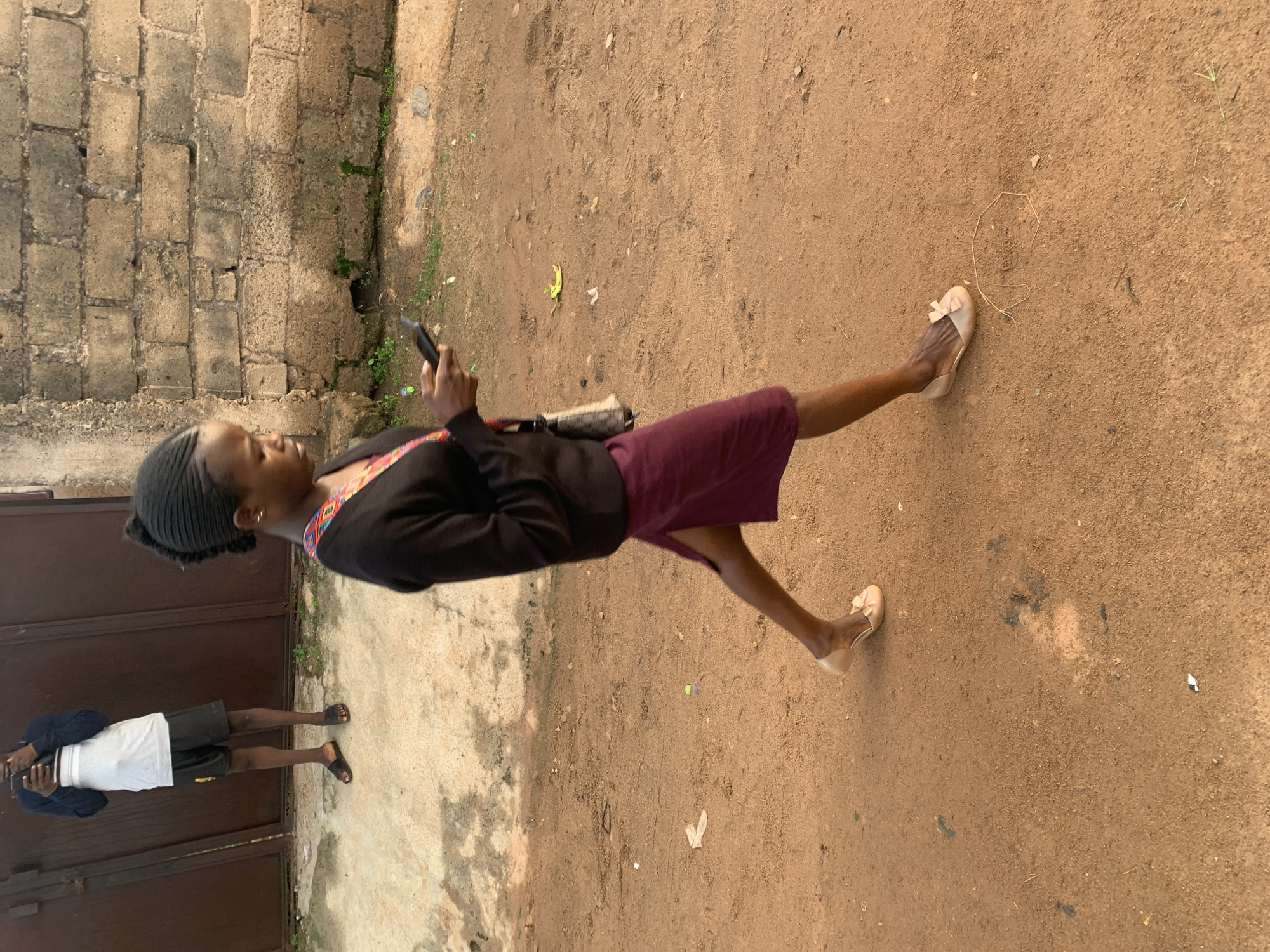 A young African woman walks on a dirt path, holding a mobile phone in her right hand. She wears a black sweater with a colorful trim around the neckline, a maroon skirt, and light-colored ballet flats with bows. Her hair is braided and pulled back. She is looking down at her phone as she walks. In the background, a man stands near a large, dark metal gate, holding a phone. He is wearing a white t-shirt with a horizontal stripe, black shorts, and black flip-flops. A brick wall is visible behind him. The scene appears to be outdoors, in a somewhat unkempt area with scattered debris on the ground, including a banana peel and some plastic wrappers. The ground is composed of reddish-brown dirt and sand. The lighting suggests it is late afternoon or early evening.